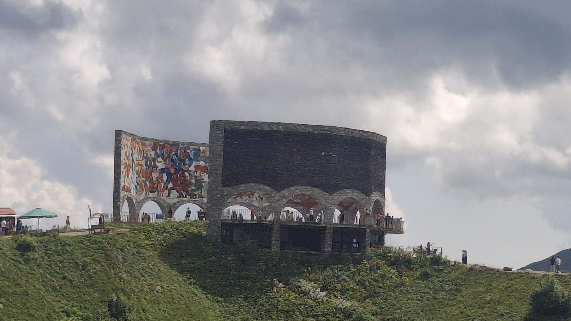 The image shows the Gudauri Friendship Monument, also known as the Georgia-Russia Friendship Monument, located in Gudauri, Georgia. The monument is a large, circular structure built on a hillside, overlooking the Caucasus Mountains. The monument features a mural on one side depicting scenes from Georgian history and mythology, while the other side is a dark, stone-faced cylindrical structure. In front of the monument, there is a walkway with railings where several people are standing and observing the view. The surrounding environment is a lush green hillside covered with vegetation, under a cloudy sky. In the foreground, there are also some small structures and umbrellas, suggesting a tourist area.