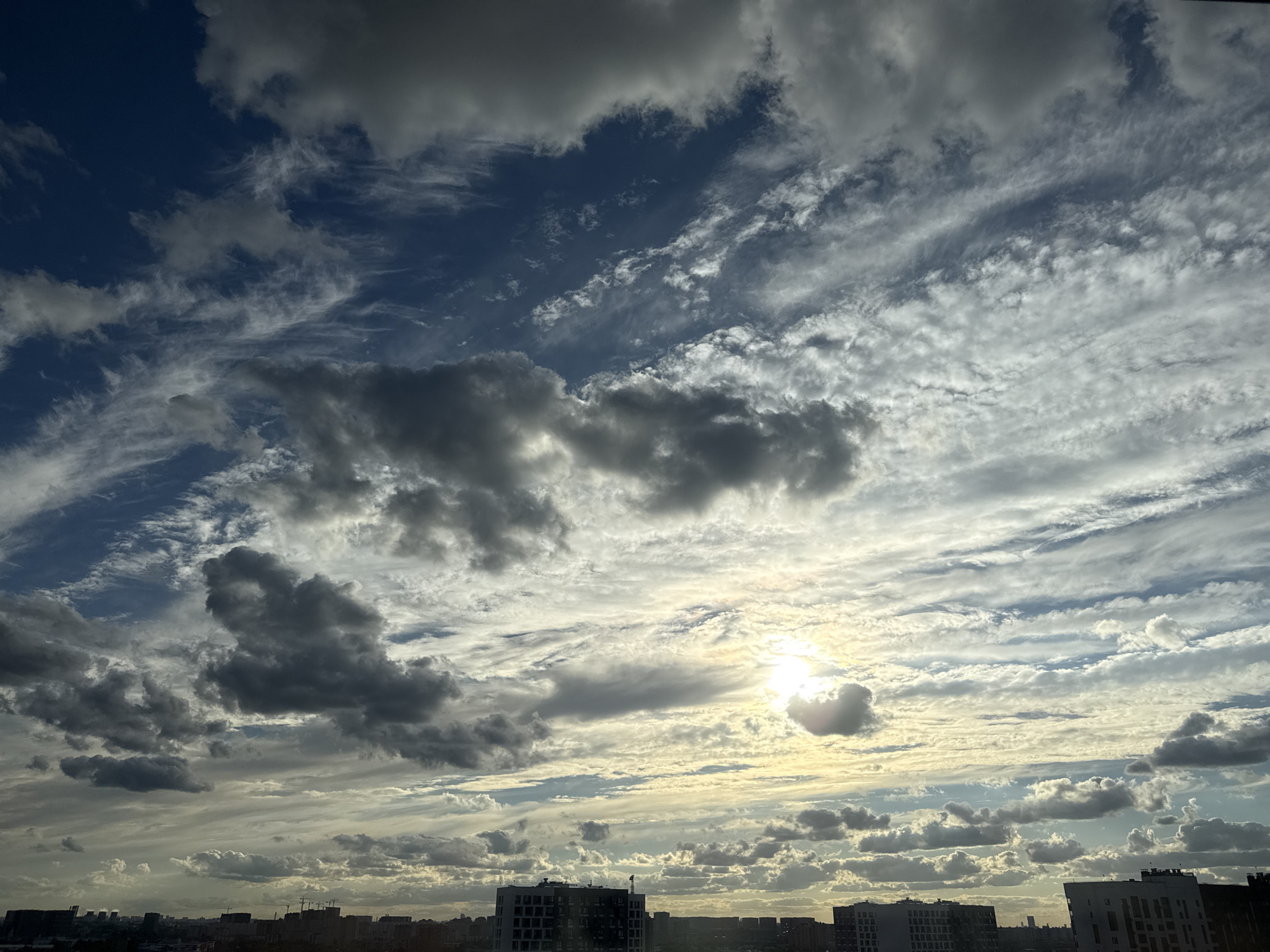A photograph taken from a high vantage point in Lyubertsy, Russia, captures a dramatic sky filled with a mix of dark, heavy clouds and wispy, illuminated clouds. The sun is partially visible, creating a bright halo effect as it peeks through the atmospheric layers. Below the sky, a city skyline is visible, characterized by various buildings of different heights and architectural styles, some of which appear to be under construction. The overall scene depicts a dynamic weather condition over an urban landscape.