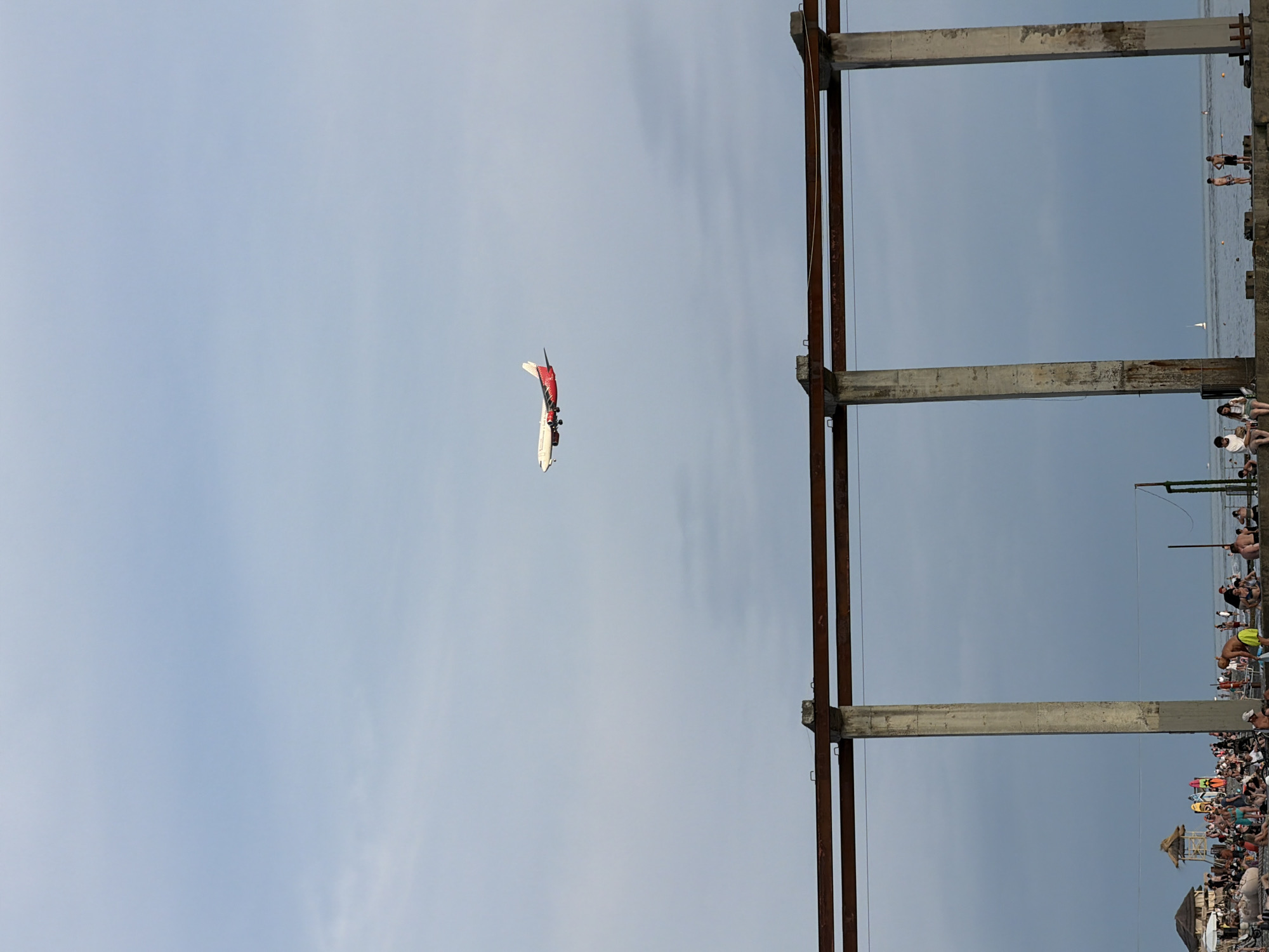 This media file depicts an aerial view of a beach scene in Sochi, Russia. The primary subjects include a crowd of people on the beach, some appearing to be swimming or enjoying the water. In the sky, a small airplane or glider is visible, decorated with red and white colors. The scene is dominated by a large, unfinished concrete and metal structure, possibly a pier or a building under construction, which frames the view of the beach and the sea. The weather appears to be overcast, with a hazy sky. The presence of the unfinished structure suggests a developing area or perhaps a relic of past construction. The overall atmosphere seems relaxed, with many people enjoying a day at the beach. The time of day is not clearly discernible but appears to be daytime.