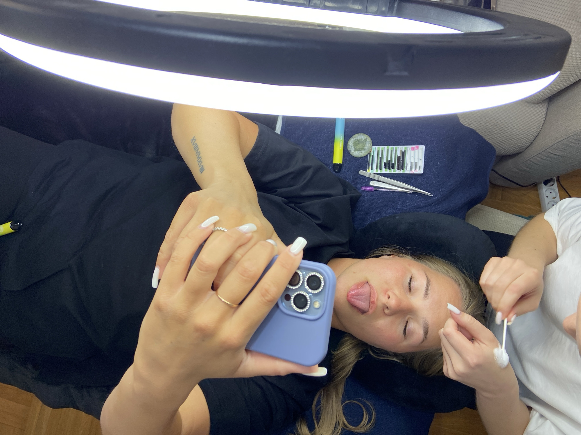 A woman lies on her back, with a bright ring light above. Another person, possibly a lash technician, is applying something to her face with a cotton swab. The woman on her back is holding a light purple iPhone up to take a selfie with her tongue slightly sticking out. On the table next to her is an e-cigarette, a glue base, and a lash palette. There are also two tweezers: one purple and one silver.
There is no indication of Budva, Montenegro in the image.