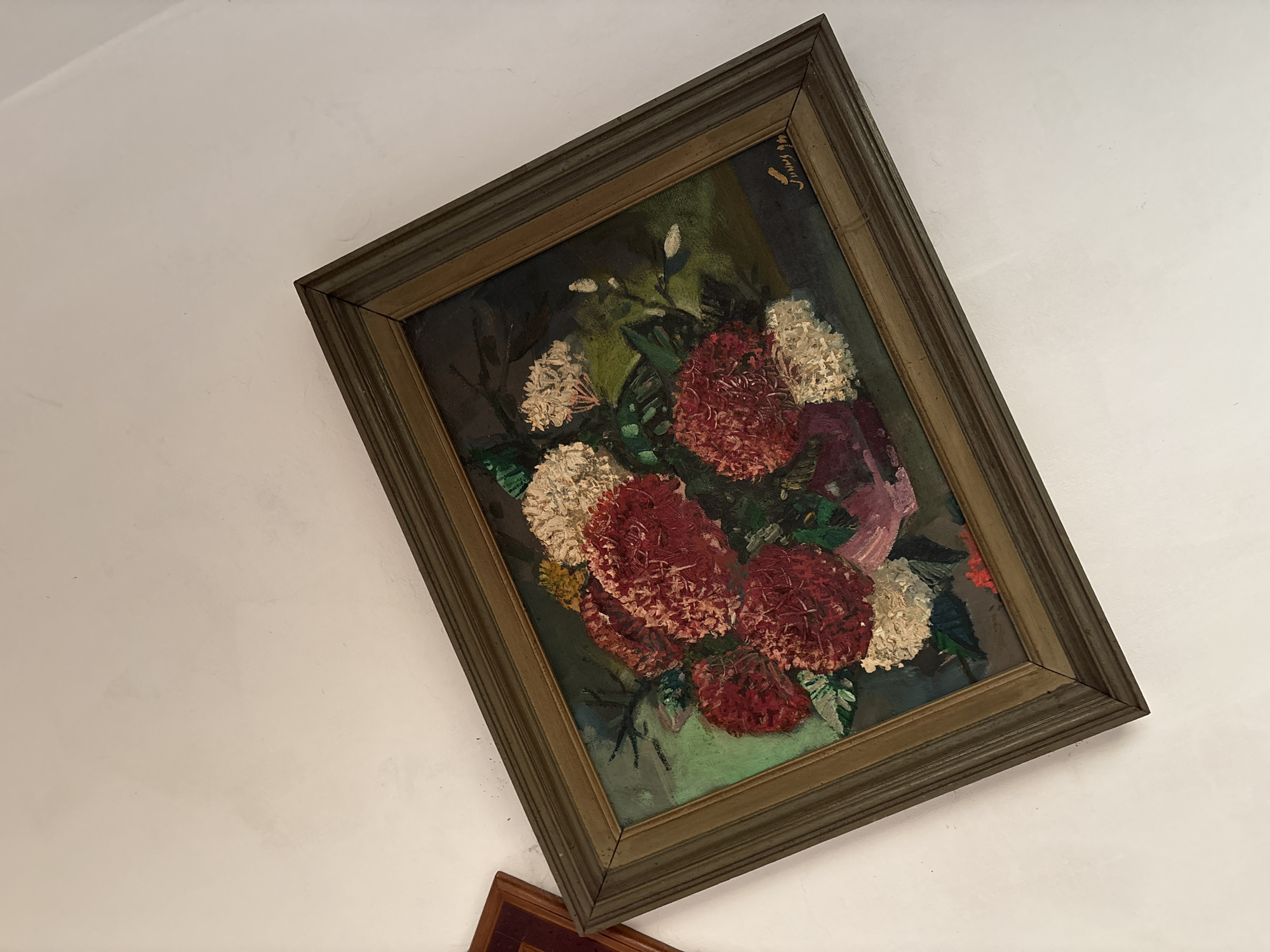 The image shows a framed oil painting of a floral arrangement with red and white flowers. The painting is hung on a white wall. The frame is made of dark wood. A portion of another framed artwork is visible on the left side of the image. The painting is signed in the lower right corner with the name "Sunny" and a date that appears to be "90". There are no people or animals visible in the image, and no context is provided regarding its location in Uleepulo, Indonesia.