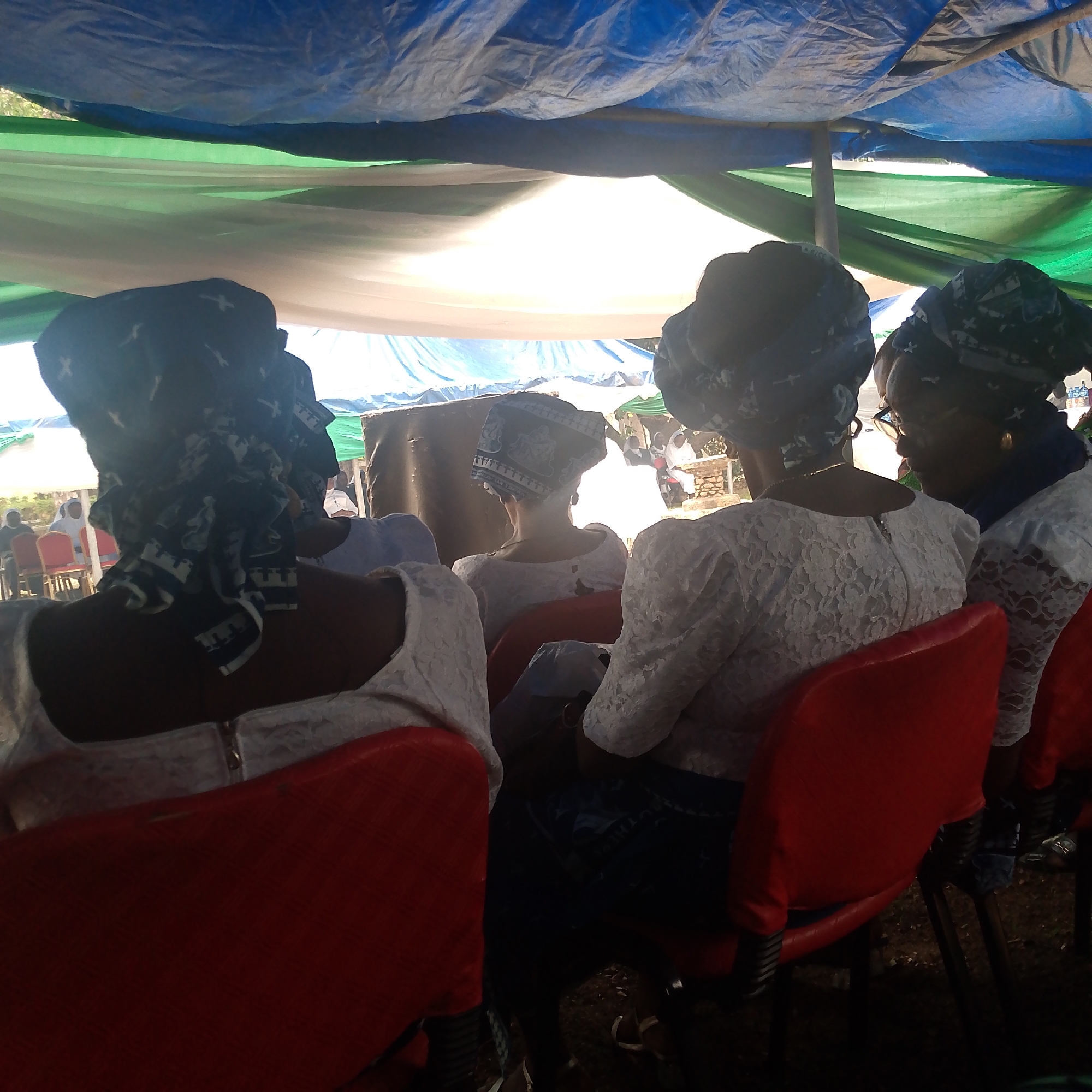 The image shows a group of women seated on red chairs under a makeshift shelter, possibly for an outdoor event. The shelter is constructed with a blue, green, and white tarp or fabric overhead. The women are wearing headwraps and white, lace-like tops. The location is in the city of Unknown, Unknown. The event may be a social gathering.