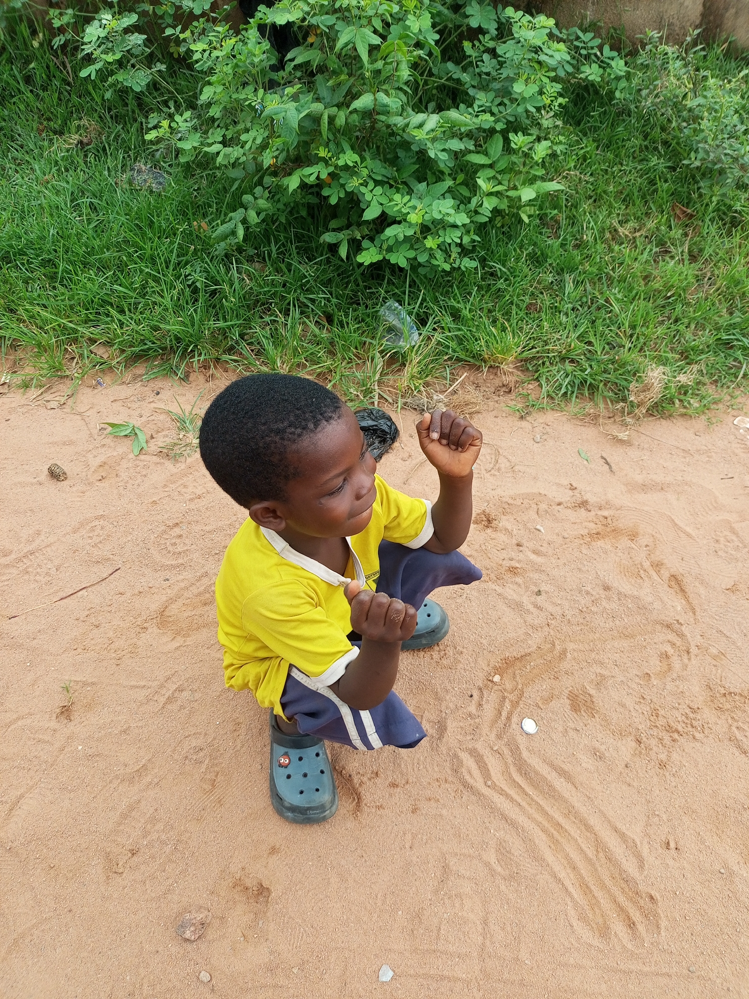 A young Black boy, appearing to be around four to six years old, is captured in a high-angle, medium shot. He is wearing a bright yellow t-shirt with a white collar and trim, and navy blue shorts with white stripes down the sides. His feet are clad in dark teal Crocs-style clogs, one of which displays a cartoonish face with prominent eyes. The boy is crouched on a sandy surface, his right hand raised in a fist as if in anticipation or excitement. He is looking upwards and to the right, with a slight smile on his face. The ground is a mixture of light brown sand with scattered pebbles and what appear to be tire tracks or indentations. Behind him, a dense patch of green grass and leafy foliage borders a rough-textured wall or building. A small white object, possibly a bottle cap, rests on the sand in the mid-ground. The scene appears to be an outdoor space, likely a yard or roadside in Gombe, Nigeria.