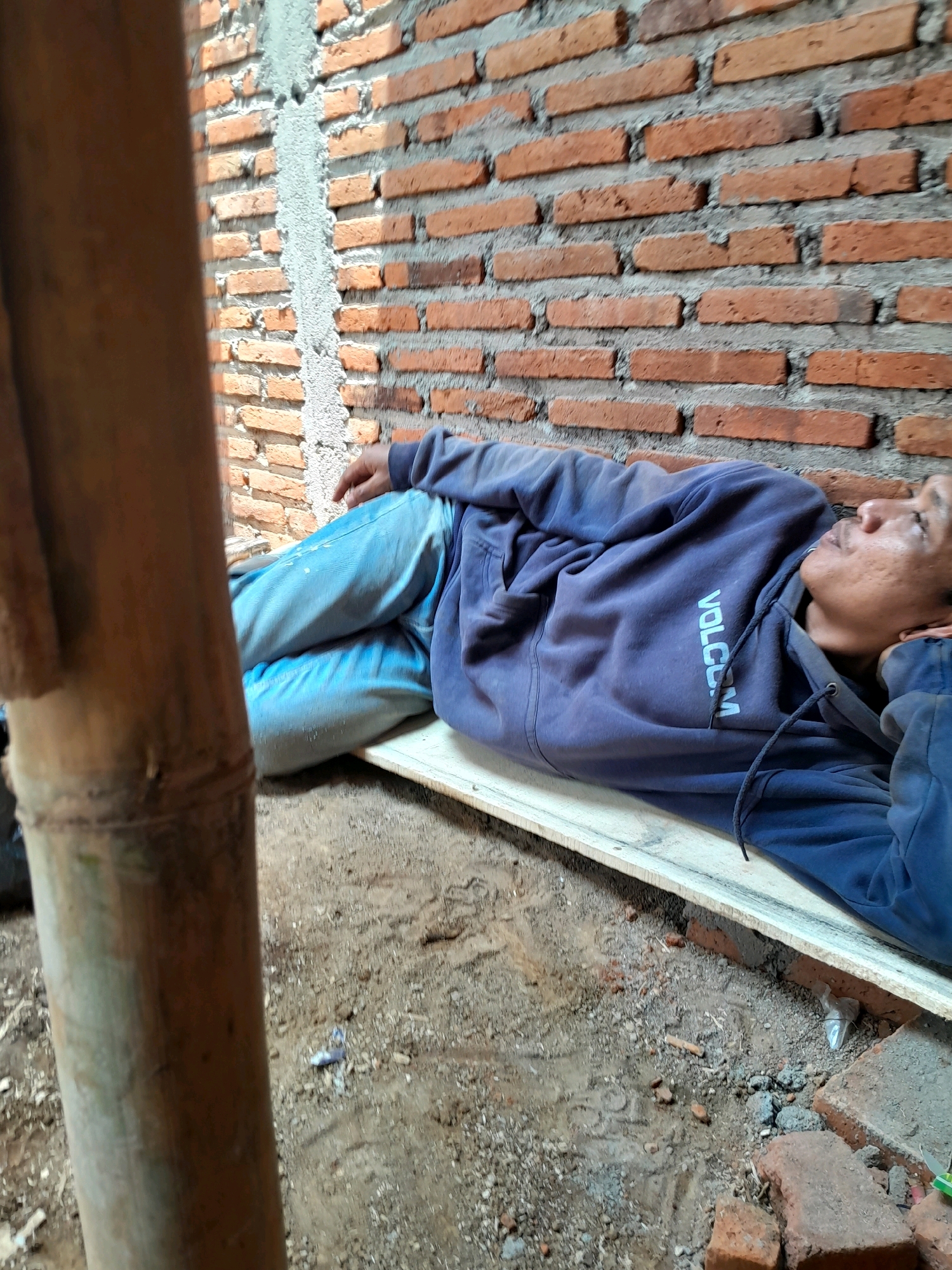 A person is lying on a wooden plank outdoors, resting against a brick wall. The person is wearing a dark blue hooded sweatshirt and light blue jeans. The environment appears to be a construction site or an outdoor area with loose dirt, bricks, and a large bamboo pole in the foreground. The brick wall is made of orange bricks with visible mortar. The lighting suggests it is daytime. The image is taken from a medium-wide angle, capturing the person and their immediate surroundings. The specific location is identified as Karangduren, Indonesia.