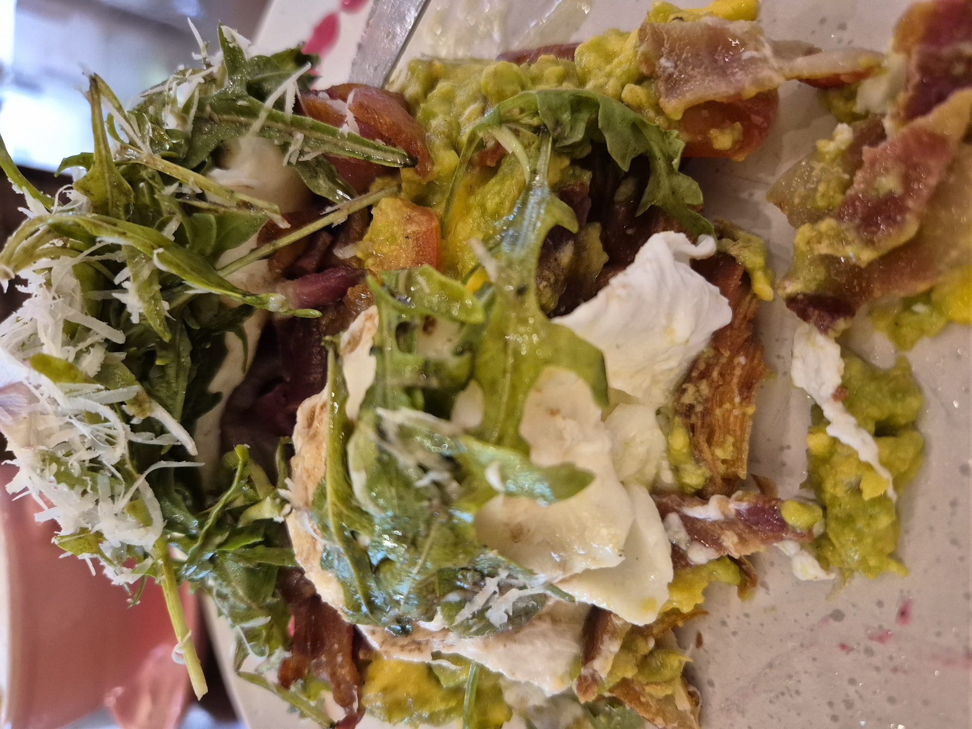 The image shows a close-up, overhead view of a dish of food, likely a breakfast or brunch item, served on a white plate. The dish appears to be an open-faced sandwich or toast topped with a variety of ingredients. Prominently featured are layers of mashed avocado, crumbled bacon, a poached or fried egg with a runny yolk, and a generous portion of arugula or similar leafy greens, possibly dressed with a light vinaigrette. Some shredded cheese, possibly Parmesan, is sprinkled over the greens. The overall presentation is somewhat messy, with ingredients spilling over the edges of the base, which appears to be a toasted bread or similar base. The colors are vibrant, with the green of the avocado and arugula contrasting with the red of the bacon and the white of the egg and cheese. The background is blurred, but suggests an indoor setting, possibly a restaurant or cafe, with a pink bowl visible in the upper left corner. There is no indication of the specific location within the city of Đà Nẵng, Vietnam.