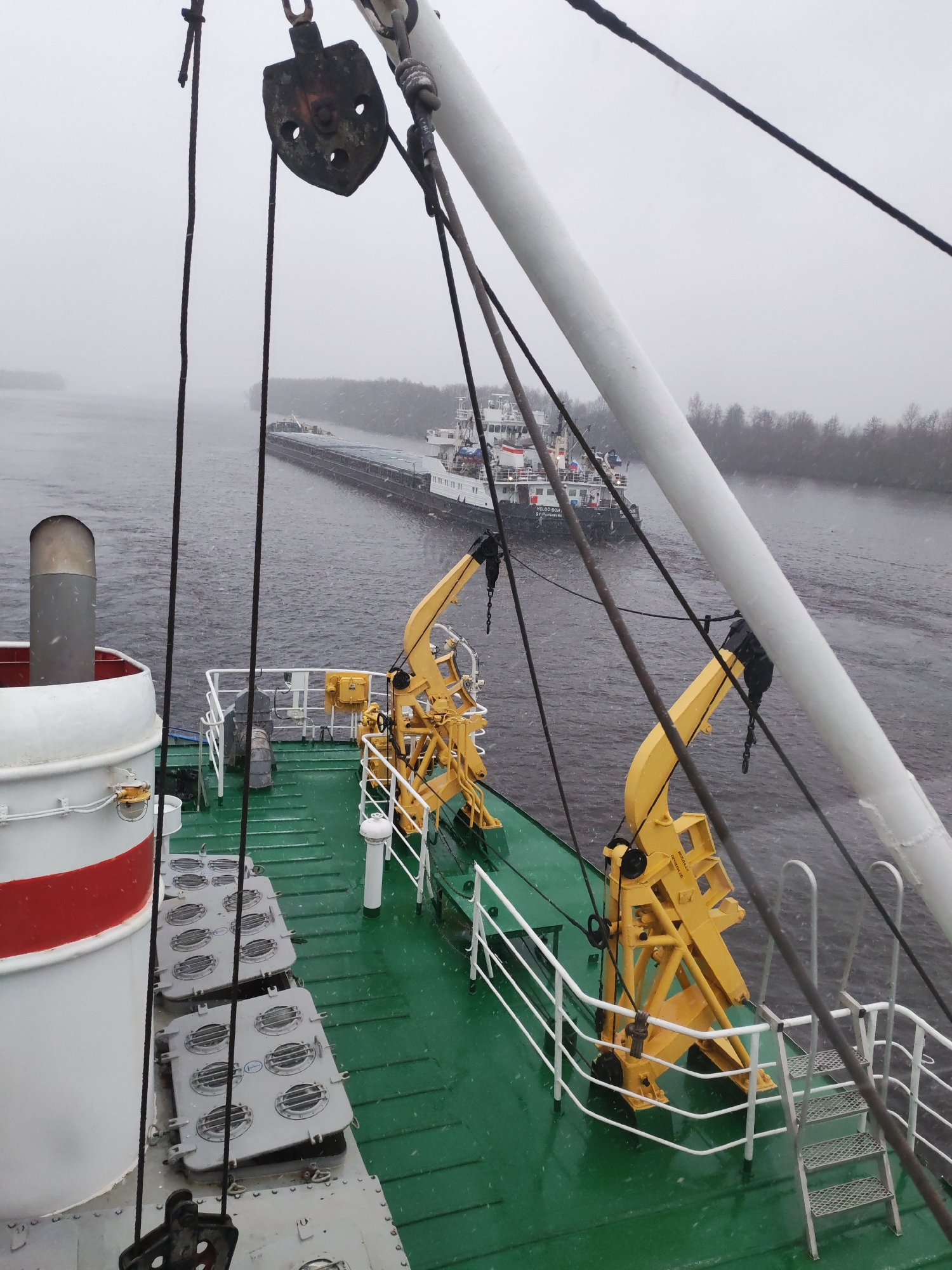 The image, taken from a boat, depicts another boat towing barges on a river in Novokemskii, Russia. Snow falls on the green deck of the boat from which the image is taken. Yellow machinery and white railings are visible. The river is grey and the sky is overcast. The opposite shore has trees without leaves.