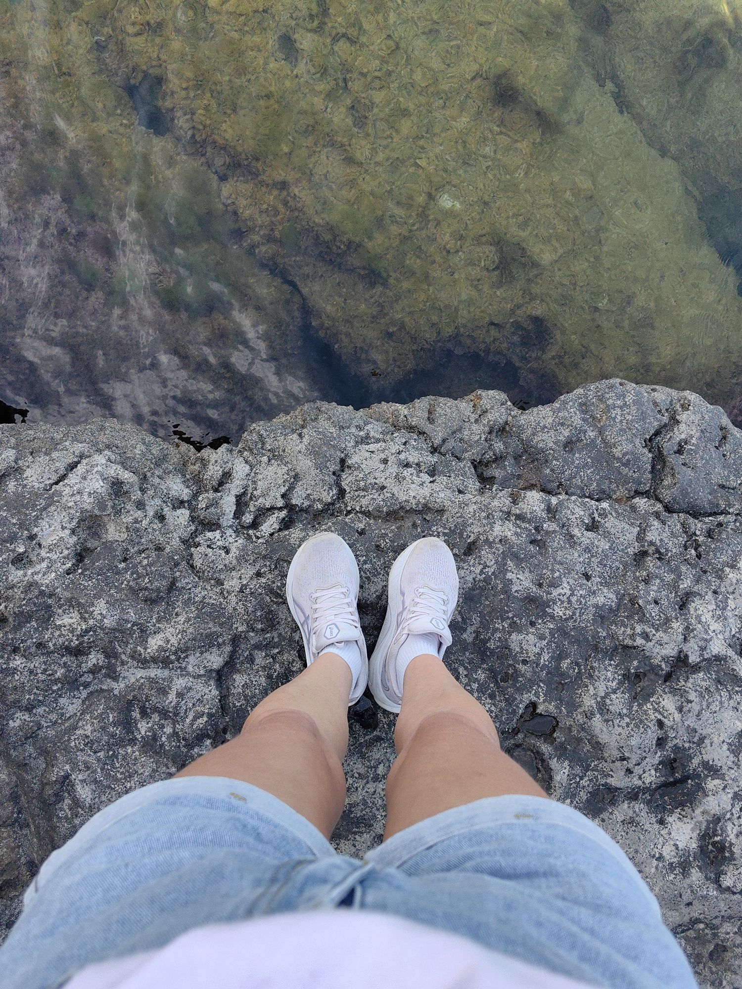 This is a top-down, eye-level medium shot of a person's legs and feet standing on a rocky surface next to a body of water. The person is wearing white athletic shoes with white socks and light blue denim shorts. Their legs are bare and tanned. The rocky surface appears rough and grey, with some patches of lighter color, possibly lichen or moss, and dark wet areas.
Beyond the rocks, the water is visible. It appears shallow and clear, with a textured bottom that shows shades of green, brown, and grey, suggesting submerged rocks or vegetation. The water's surface reflects light, creating a shimmering effect. The overall scene suggests an outdoor, natural setting, possibly a coastline or a rocky lake shore.
The lighting appears to be natural daylight, casting subtle shadows and highlighting the textures of the rocks and water. There is no explicit indication of the time of day or weather conditions beyond it being daylight. The composition focuses on the ground and the lower extremities of the person, giving a sense of standing at the edge of something. There is no visible text in the image. The scene evokes a feeling of contemplation or a pause during an outdoor activity.