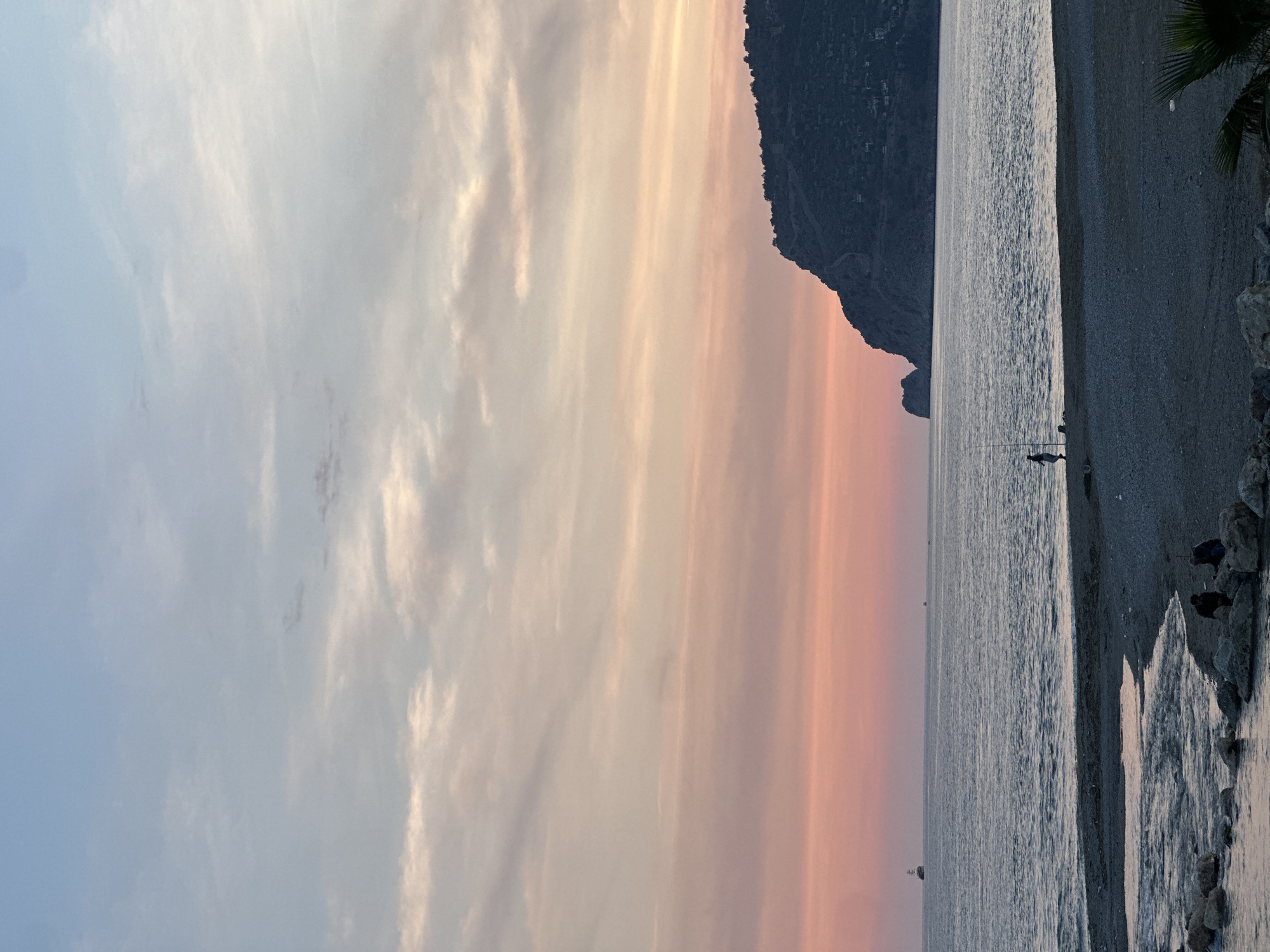 A photograph shows a coastline at sunset. The sky transitions from pale blue on the left to a warm orange/pink hue towards the right. Wispy clouds are scattered across the sky.
On the right side of the frame, a dark, mountainous headland juts out into the water. The sea is calm, with gentle ripples and reflections of the sunset colors.
The coastline is a pebble beach with rocks and at least three people are seated on the rocks. Two figures, seemingly fishermen, are visible in the distance, standing near the water's edge.
The location is Alanya, Turkey, as implied by the terrain and coastal setting.