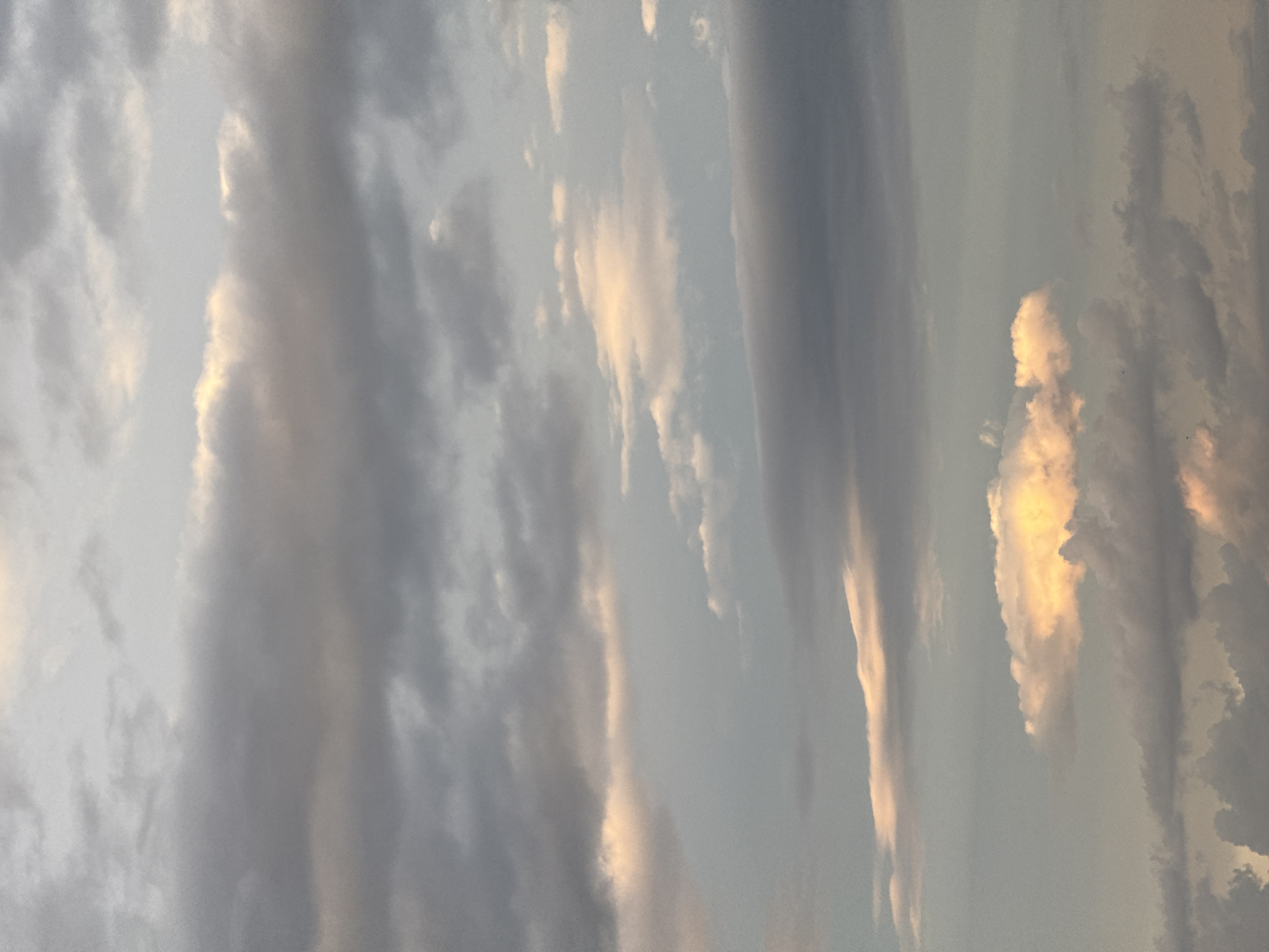 The image shows a sky with streaky, layered clouds. Some clouds are highlighted with a golden or yellow light, likely from the sun. The overall color palette consists of gray, blue, and golden tones. The scene appears to be in the city of Unknown, Unknown. No people or objects are visible other than the sky and clouds.