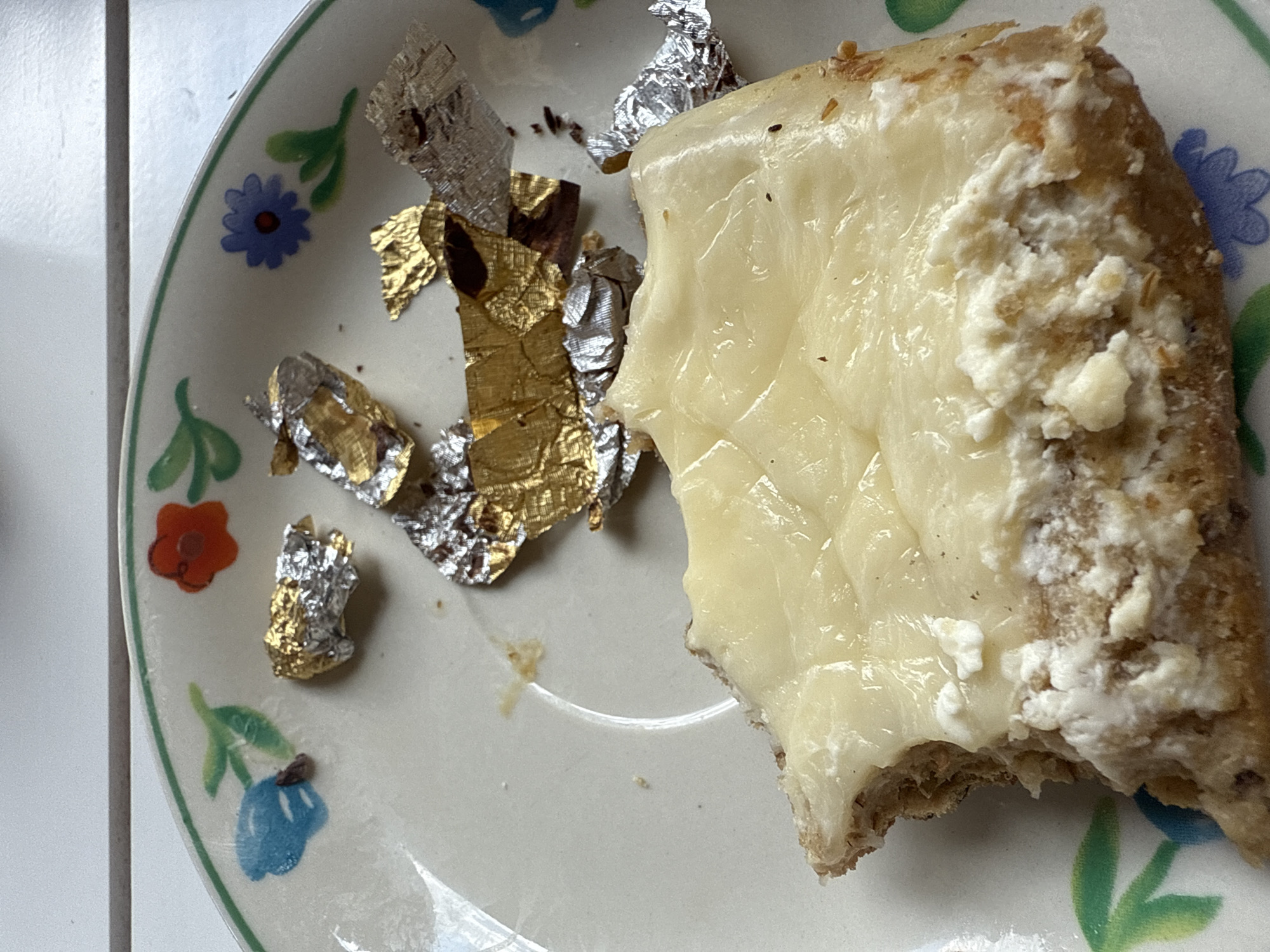 A slice of bread with a creamy white topping is on a plate with a floral design. There are pieces of gold and silver foil scattered on the plate. The plate is white with green trim and painted blue and orange flowers.