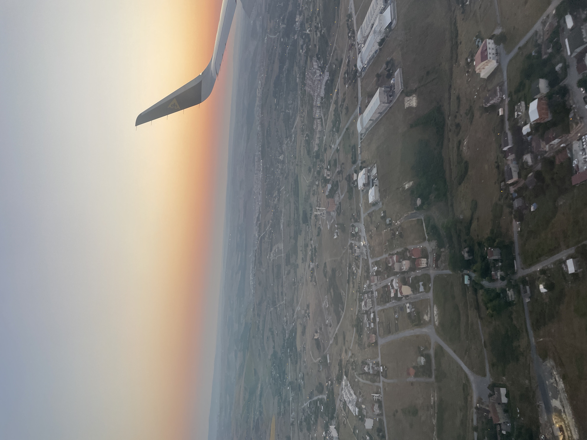 An aerial view captures a suburban landscape at dusk, possibly in Beykoz, Turkey, with a prominent airplane wing in the upper right quadrant. The sky transitions from a pale blue to a soft orange and pink hue, indicating sunset. Below, a patchwork of residential buildings, some with red or beige roofs, are scattered amongst fields and undeveloped land. Roads wind through the area, connecting the houses and leading towards larger structures resembling warehouses or industrial buildings visible on the right side. The overall scene conveys a sense of calm as day transitions into night, with the airplane suggesting travel or arrival.