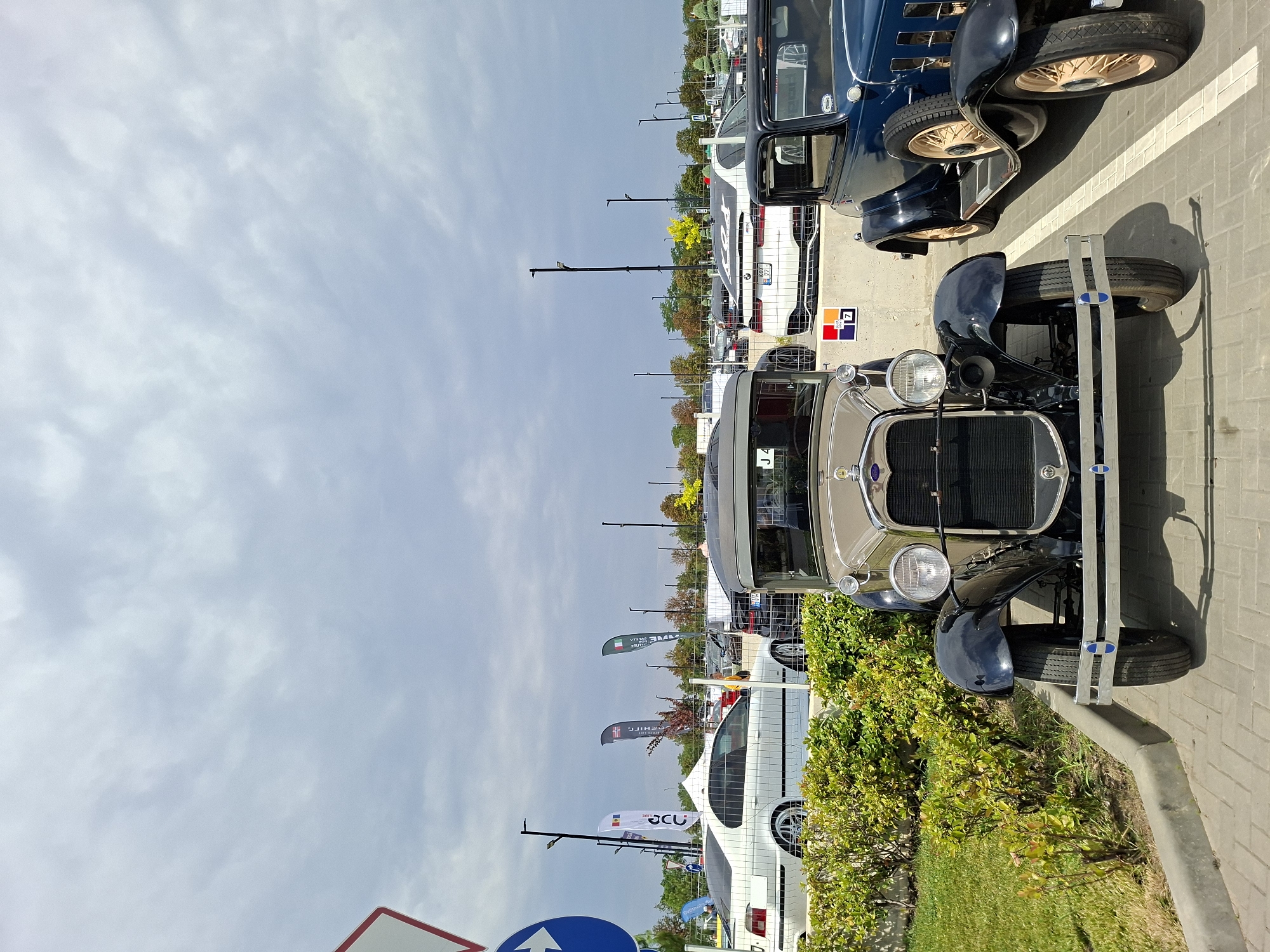 The image depicts an outdoor event, likely a car show, in Stăuceni, Moldova. The foreground features two vintage cars. The car on the left is a tan and black sedan, and the car on the right is a dark blue sedan with tan wheels. Both cars are parked on a paved surface in front of a low concrete barrier and a green hedge. In the background, several modern cars are visible, along with flags and tall poles with lights. A white sedan is parked to the left of the vintage cars, behind a fence. The sky is overcast. There are no people clearly visible in the image. The scene appears to be a sunny day with cloudy skies.