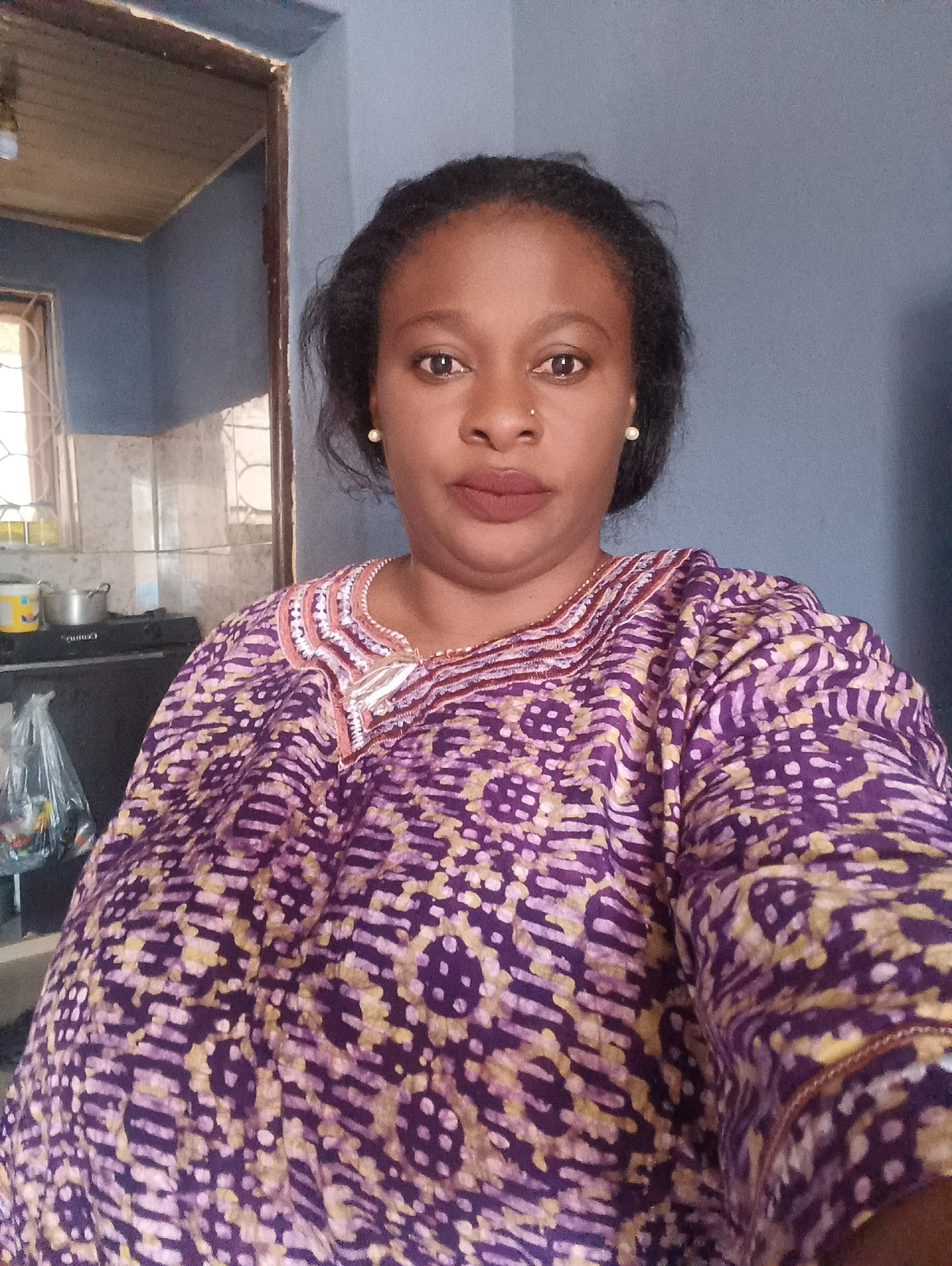 A woman is standing indoors, possibly in a kitchen, in Jos, Nigeria. She is wearing a purple and yellow patterned top with decorative trim around the neckline. She has dark hair pulled back, is wearing small earrings, and has a nose piercing. The background shows a section of a room with blue walls, a window, and kitchen appliances. A stove, pots, and a bag of food are visible.