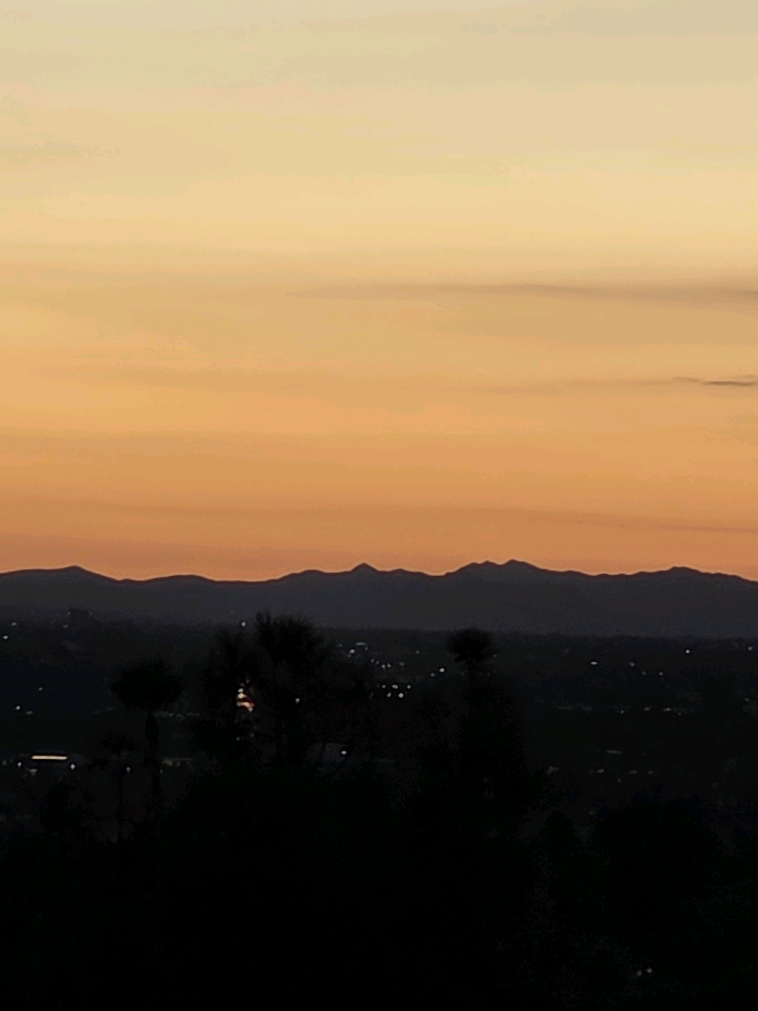 This is a landscape photograph taken at dusk in Glendale, United States of America. The scene depicts a silhouette of mountains against a sky filled with gradient hues of orange and yellow, indicative of a sunset. In the foreground, dark silhouettes of trees and possibly some structures of a cityscape are visible against the darkening terrain. The overall impression is a peaceful evening view of a natural landscape bordering a populated area.