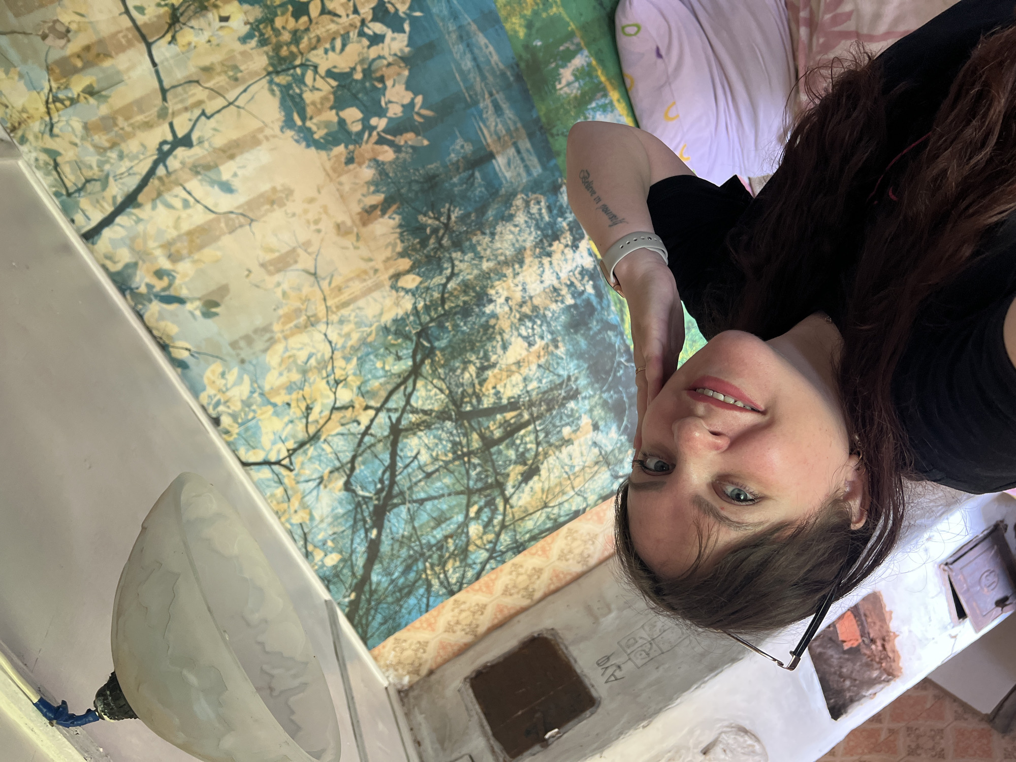 The image is a portrait of a woman, taken from a low angle. She is looking at the camera with a slight smile. She has long, dark brown hair and is wearing a black t-shirt. There is a tattoo on her left forearm that reads "Believe in yourself". She is wearing a silver fitness tracker on her left wrist.
The background is a colorful mural depicting trees with light-colored leaves against a backdrop of blue and green. To the right of the mural, there are pillows with a pink and white floral pattern.
The woman appears to be in an indoor setting, possibly a bedroom or a room with a decorative mural. There is a white lampshade on the left side of the image, and a small, rustic stove or hearth in the lower right corner. The lighting suggests it is daytime. The overall mood is casual and relaxed.