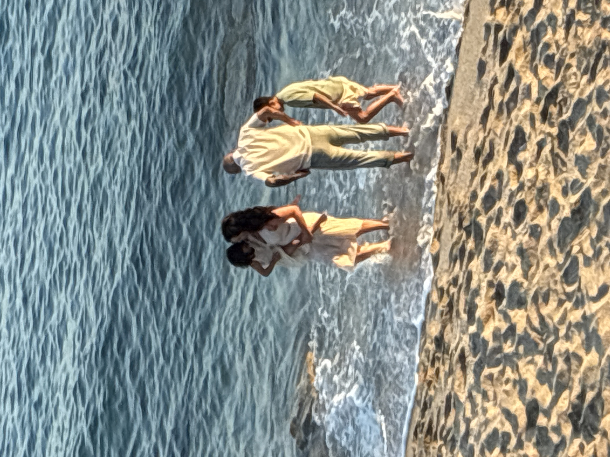 The image shows three people at the edge of the sea in Alanya, Turkey. The scene is oriented vertically, with the sea on the left and a textured, stony embankment on the right. The water is a vibrant blue with visible ripples and white foam from the waves. A person in a light-colored shirt and pants is standing in the water, facing away from the camera, with their arm raised. They appear to be interacting with another person, also in light-colored clothing, who is partially submerged in the water and facing towards the standing person. A third person, dressed similarly, is positioned lower in the frame, also in the water, with their head turned.
The lighting suggests it is daytime, possibly late afternoon given the warm tones on the embankment. The activity appears to be a playful interaction or pose in the shallow water near the shore, with the waves gently lapping around them. The embankment on the right side of the image has a rough, uneven texture with variations in color, likely representing natural rock or a man-made structure using local stone. The overall mood seems relaxed and serene. There is no visible text in the image.