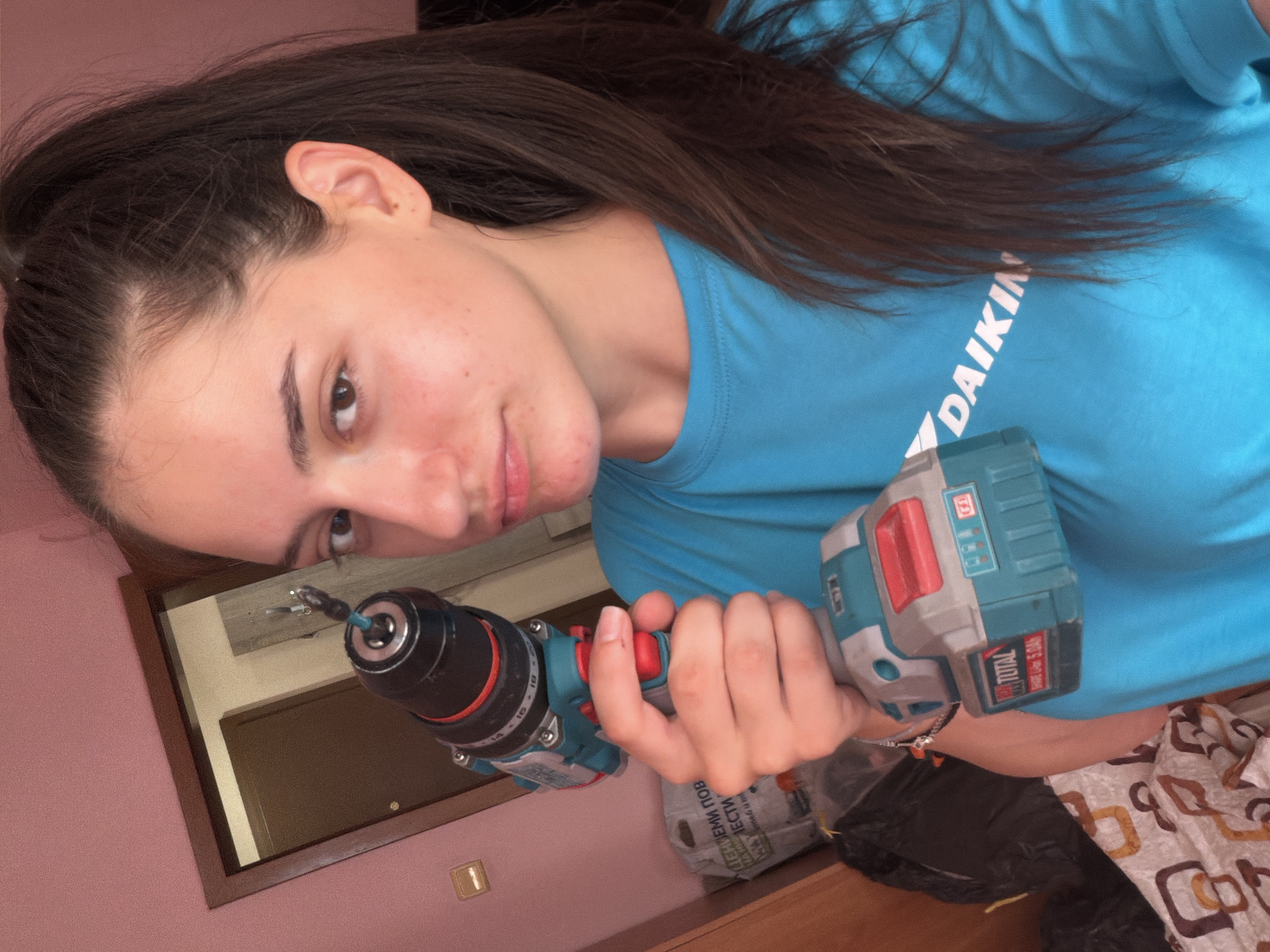 A young woman with dark hair tied in a high ponytail is holding a cordless drill in her right hand, bringing it up towards her face as she looks into the camera. She is wearing a bright blue t-shirt with a white "DAIKIN" logo visible on the right side of her chest. The drill is turquoise and gray with red accents, and a battery pack labeled "TOTAL SHARE Li-ION 5.0Ah" is attached to the bottom. Her left hand is not fully visible but appears to be holding the drill body. The background consists of a pink wall, a doorway with a dark wooden frame revealing a glimpse of another room, and various objects including bags and bedding. The scene suggests an indoor setting, potentially a home, possibly in Pomorie, Bulgaria, given the context.