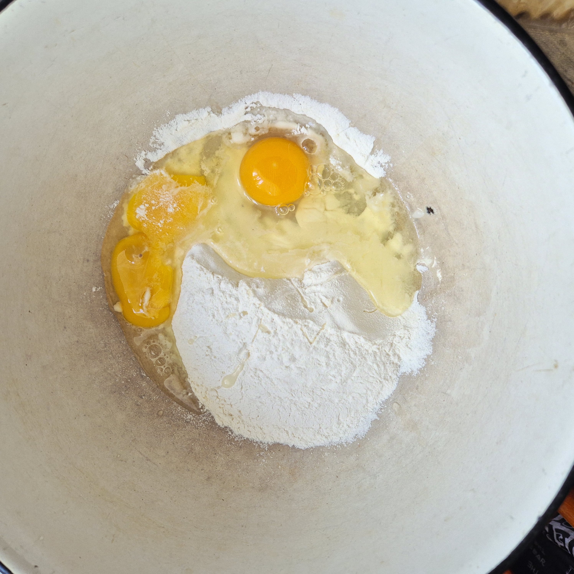 This is a top-down, medium shot of a light-colored mixing bowl containing flour and two raw eggs. The flour is piled on the left side of the bowl, and the two eggs, one whole with its yolk intact and another broken with the yolk and some white visible, are on the right side. The bowl appears to be made of ceramic or enamel and has some scuff marks. The background is blurred and appears to be a wooden surface. The location of this scene in Liski, Russia cannot be determined from the provided image.