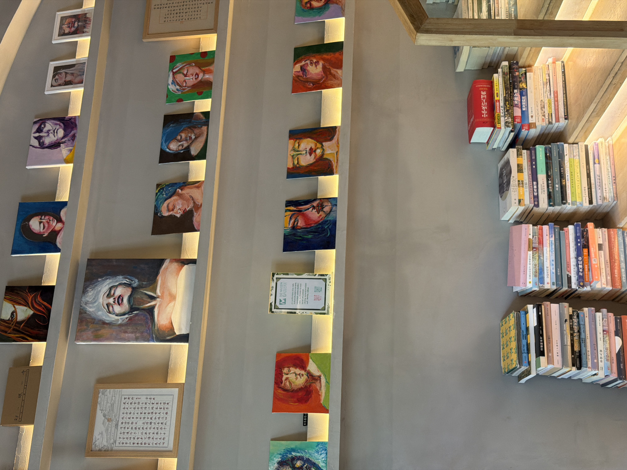 This image shows a wall adorned with paintings and bookshelves. On the left, vertically arranged paintings of faces line the wall, illuminated by built-in lighting strips. A framed text document is present on the lower-left. To the right, bookshelves are filled with various books, neatly arranged. The overall color scheme is muted, with grays and neutral tones dominating the scene. The location is within the Dongcheng District of China, as hinted by the text visible within the image.