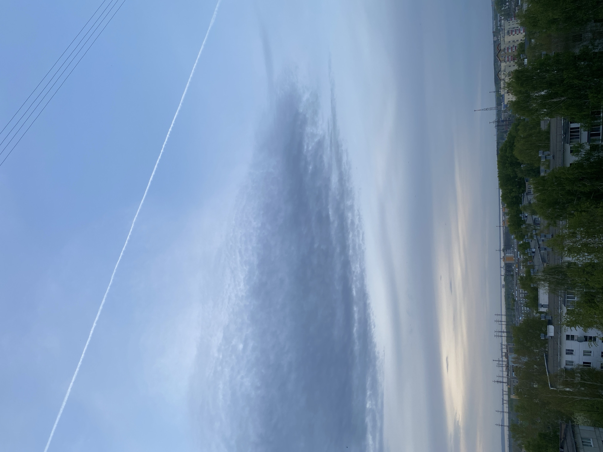 The image shows an urban landscape under a cloudy sky. In the foreground, power lines are visible against the sky, along with a contrail from an aircraft. The middle ground contains a dense cloud formation. The background shows a cityscape with buildings, trees, and industrial structures, including what appears to be a construction crane. Based on the presence of the power lines and the urban architecture, it is likely that this image was taken in an area near residential or industrial zones. Without additional information such as street names or unique landmarks, identifying the specific location within Chaikovsky, Russia is not possible.