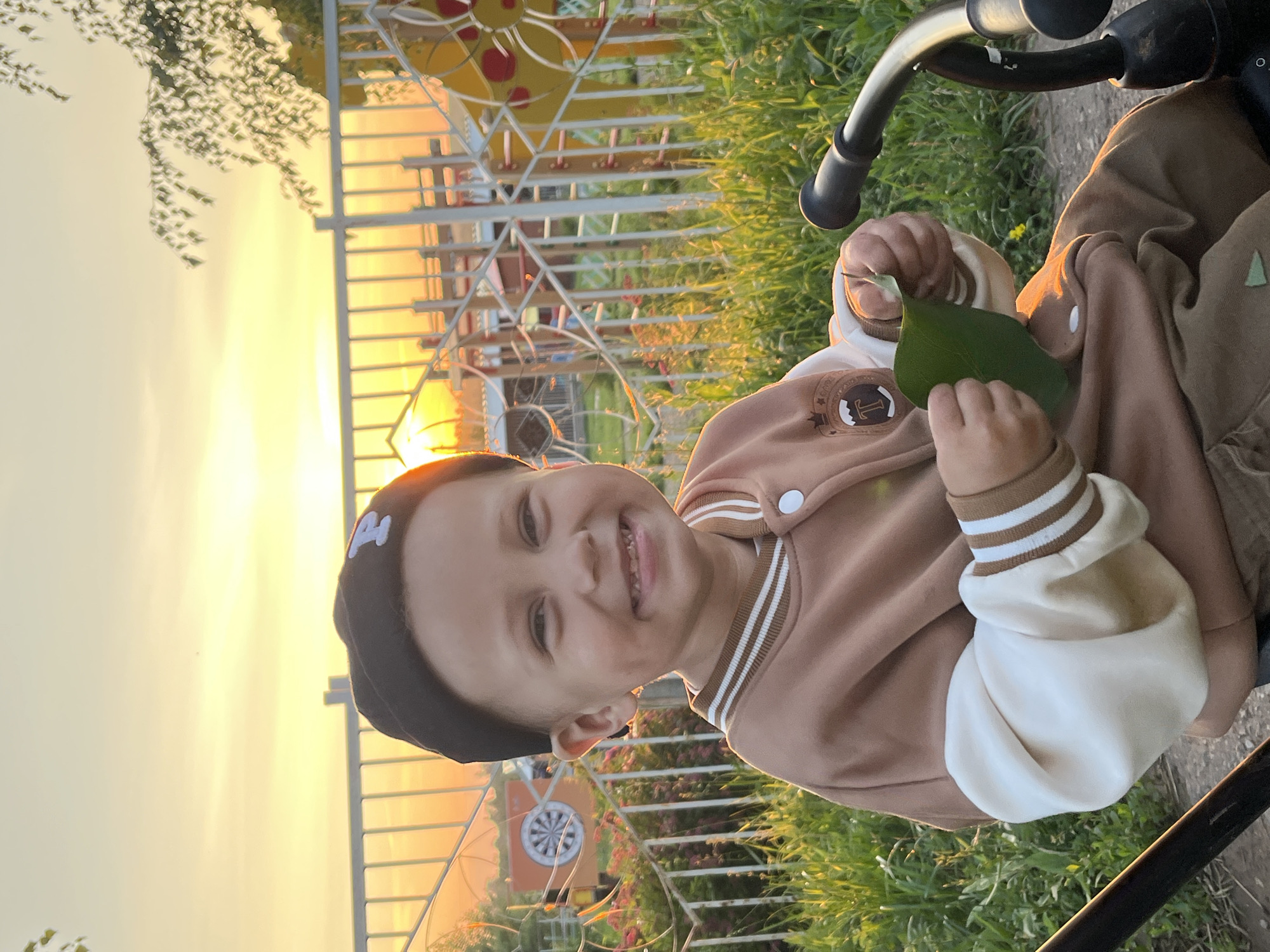 In the city of Fokino, Russia, at sunset, a young boy sits on a bicycle. He is wearing a black beanie with a white 'P' on it and a brown and white bomber jacket. The boy is smiling and holding a green leaf. In the background, a white fence surrounds a playground. The sun is setting, casting a warm glow on the scene.