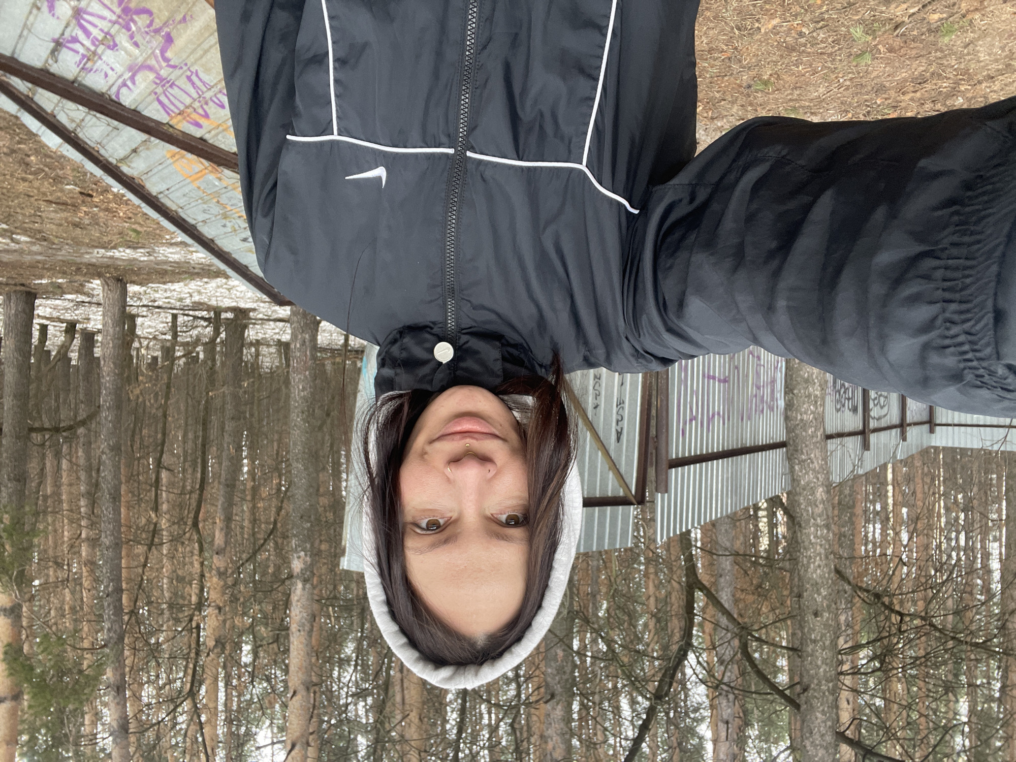 A woman with dark hair is wearing a hooded sweatshirt with a white hood, and a black Nike jacket with white accents. She is outdoors in a forest with tall, thin trees. There is a corrugated metal structure with graffiti visible in the background. The location is Izhevsk, Russia.