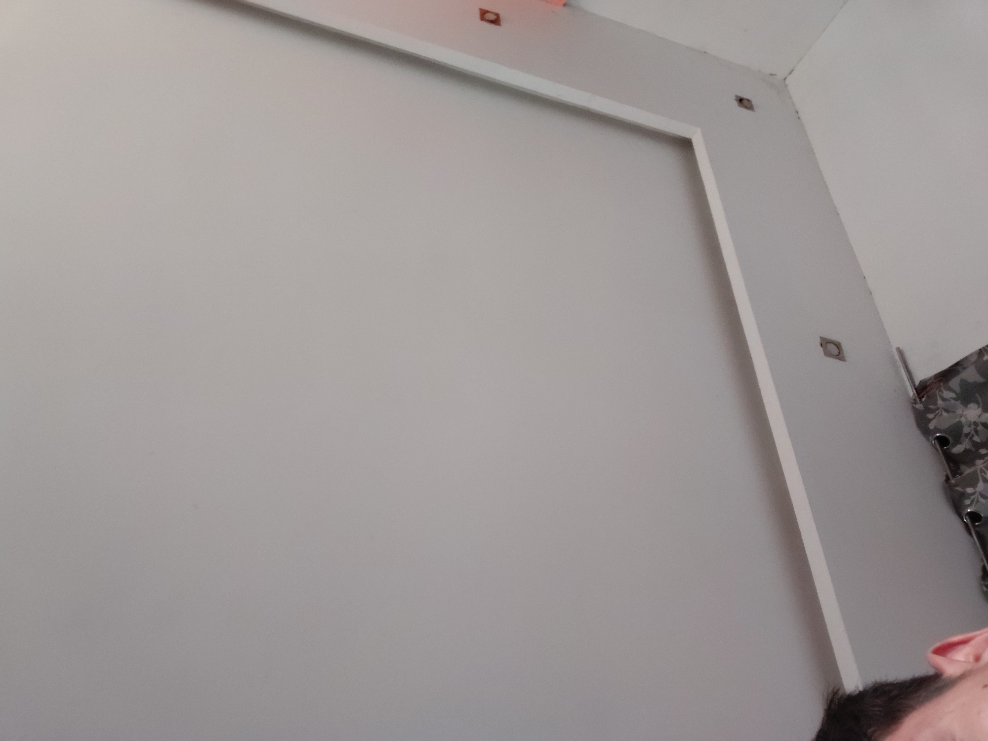 The image displays an indoor scene, possibly a room in Santa Ana, Venezuela, likely under construction or renovation. The majority of the frame is dominated by a plain, light gray wall. A white trim or frame outlines a rectangular section on this wall, forming an "L" shape with its horizontal segment along the top and vertical segment extending downwards on the right.
Embedded within this white trim and the adjacent wall sections are several small, square-shaped openings or electrical boxes. Two of these are visible along the top horizontal trim, emitting a faint orange or reddish glow, suggesting indicator lights or active electrical components. Another similar cutout is located higher up on the vertical white trim, and a fourth is visible further down on the wall section to the right of the trim.
In the bottom right corner, partially cropped, the top of a person's head is visible, showing dark hair, a forehead, and part of an ear. To the far right, a piece of patterned fabric, dark in color with lighter floral or leaf designs and visible silver grommets or rings, hangs vertically, possibly a curtain or tapestry. No specific activity is clearly depicted, but the exposed electrical elements suggest work in progress. The lighting appears uniform, and there is no visible text in the image.