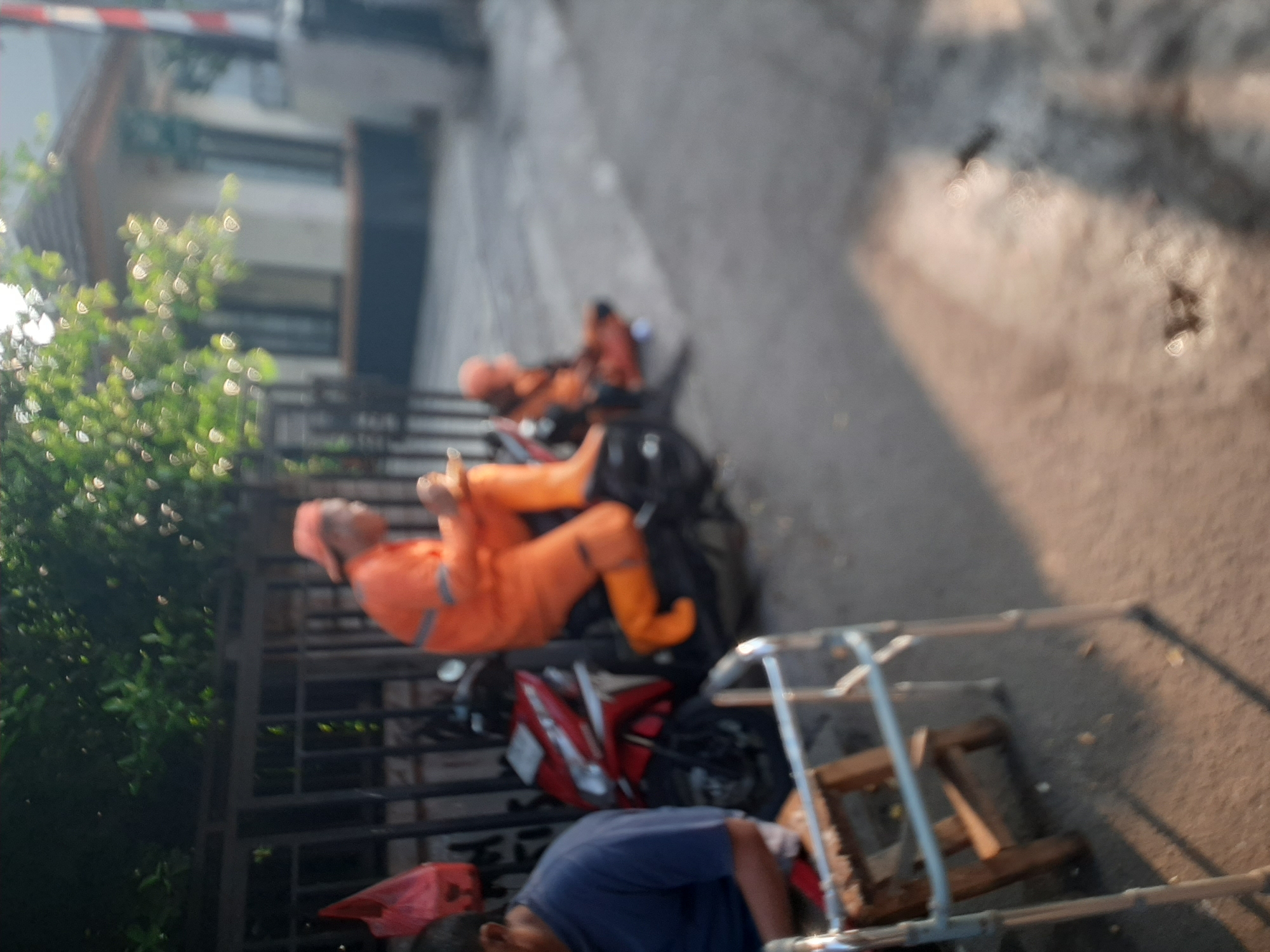 The image shows a street scene with several figures and objects. In the foreground, a metal walker is positioned on the asphalt. To the left, a person wearing a blue shirt is seated, facing away from the camera. Further back, a person in an orange uniform, including a hat and bright orange overalls, is seated on a red motorcycle. This individual appears to be holding a small object. Behind the motorcycle, another person in a similar orange uniform is seated on the ground, also appearing to be looking down. A red plastic bag is hanging on a fence in the background. A building with windows and a tiled roof is visible behind the figures, along with trees and foliage. The lighting suggests it is daytime, with sunlight casting shadows. The scene appears to be an outdoor area in a residential or commercial district, likely in a city.