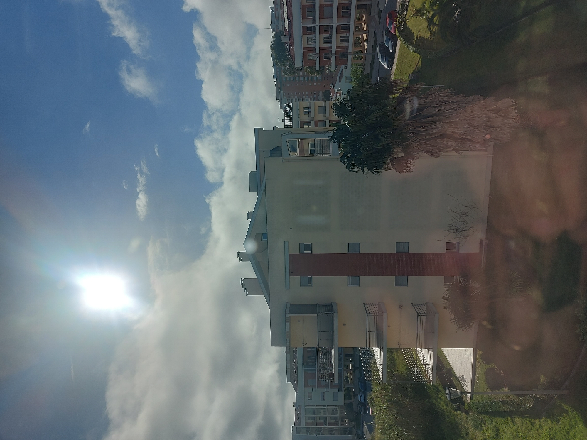 The image shows an upward-tilted view of several buildings in Figueira da Foz, Portugal under a bright, slightly cloudy sky. The sun is visible in the upper left portion of the frame. A multi-story building with a white facade and a red brick stripe runs through the center of the image. To the right is green vegetation and a blurry reflection in the window from which the picture was taken. In the background are several other buildings of varying heights and colors. Parked cars are visible along a street.