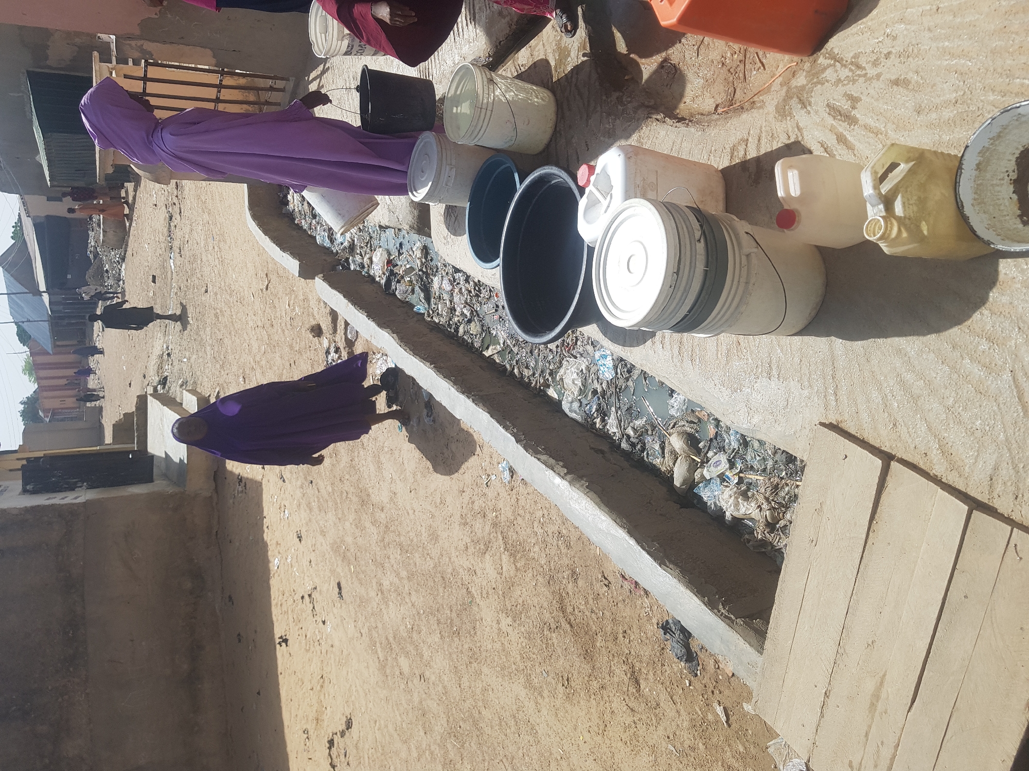 This image captures a street scene in Dutsinma B, Nigeria. Two people in purple hijabs are visible. Several plastic containers and buckets line a concrete drainage ditch filled with trash and standing water. Other people can be seen walking in the background. The buildings are low and constructed of concrete. The street is dirt.