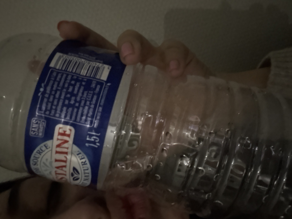 A hand with light pink nail polish holds a 1.5-liter bottle of Cristalina natural spring water. The bottle has a blue label with the brand name in red and white, alongside information in smaller text. The bottle is clear and condensation droplets are visible. The background is a dim, slightly blurred neutral tone. A portion of a sleeve with a knitted texture appears on the side.