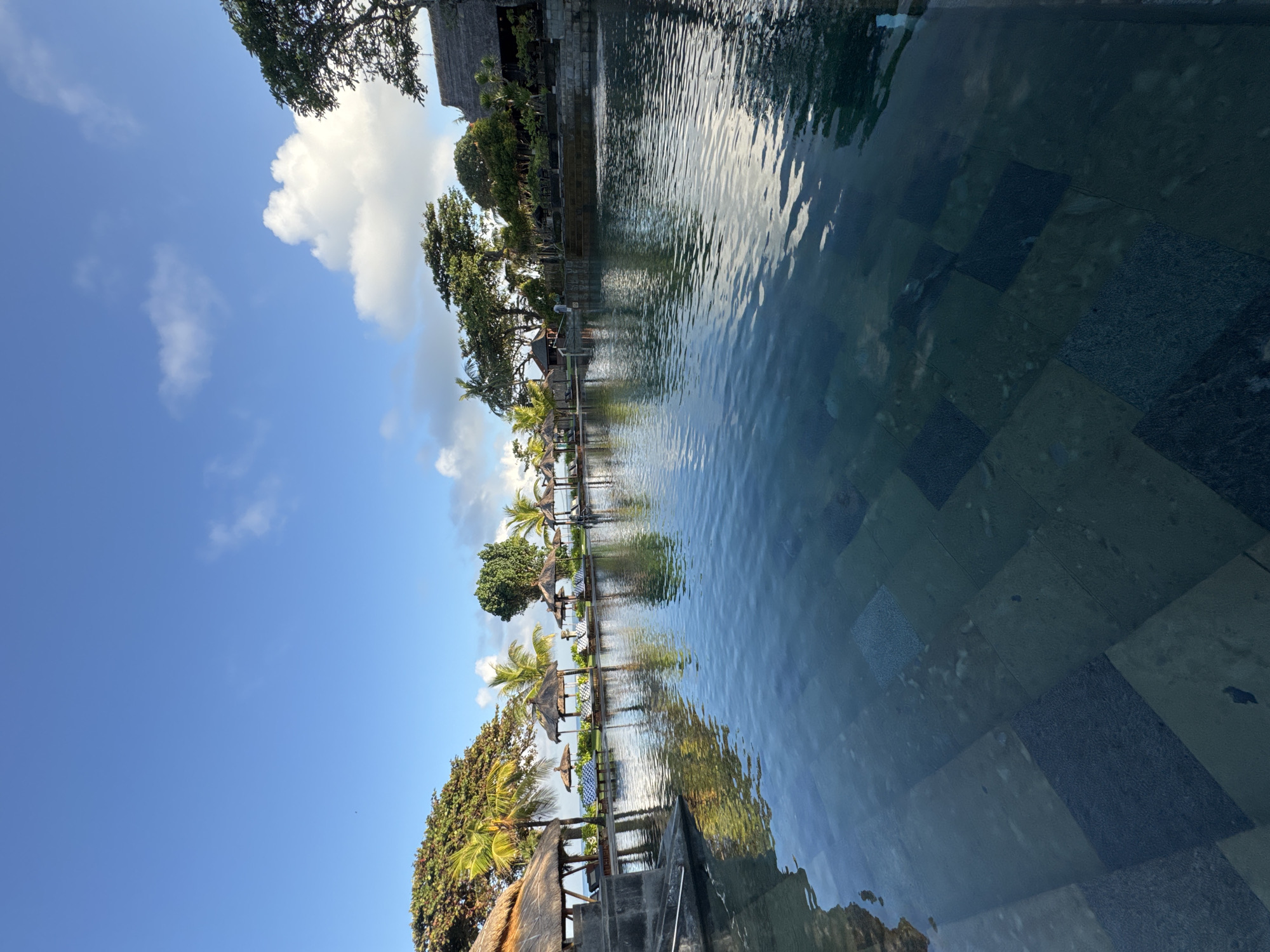 The image depicts a view from inside a pool at what is likely a resort in Jimbaran, Indonesia. The pool's water is clear with visible reflections of the sky and surrounding trees. The bottom of the pool appears to be tiled with dark-colored stone. The foreground features a set of stone stairs leading into the pool.
Beyond the pool's edge, there is a row of thatched-roof sun umbrellas with chairs and greenery between. Trees line the perimeter, with a view of a blue sky with white clouds.