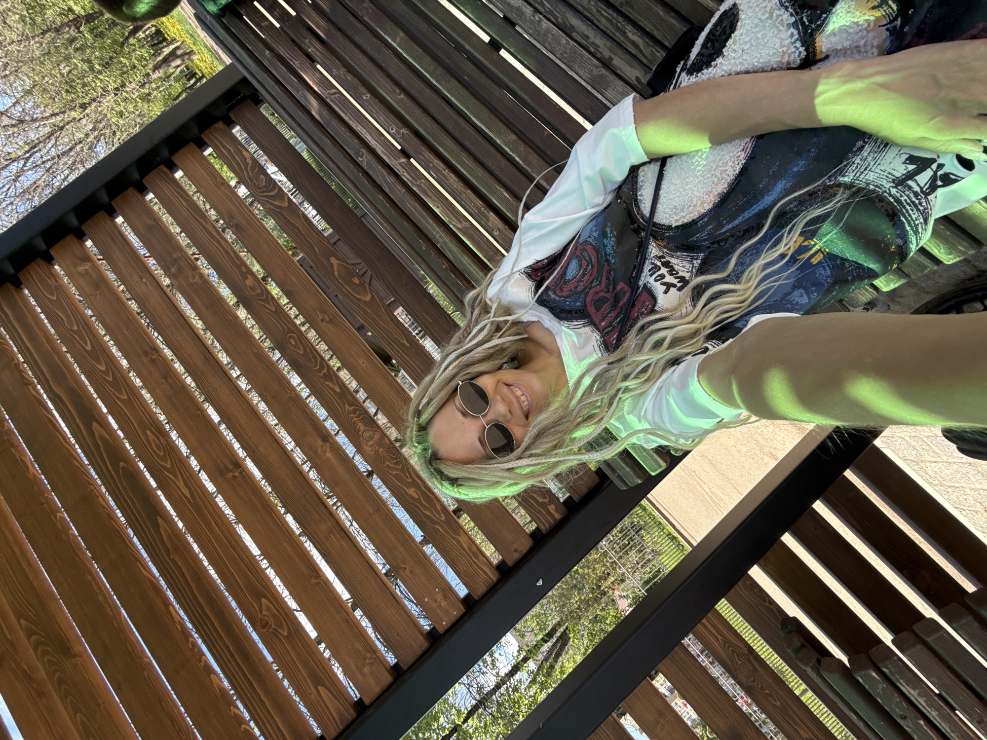 A woman with blonde dreadlocks and sunglasses is lying on a wooden structure, possibly a bench or pergola, in an outdoor setting. She is wearing a white shirt with a graphic design on the front. The background includes trees, sky, and other greenery, suggesting a park or garden. Based on the prompt, the location is Rusanovo, Russia.