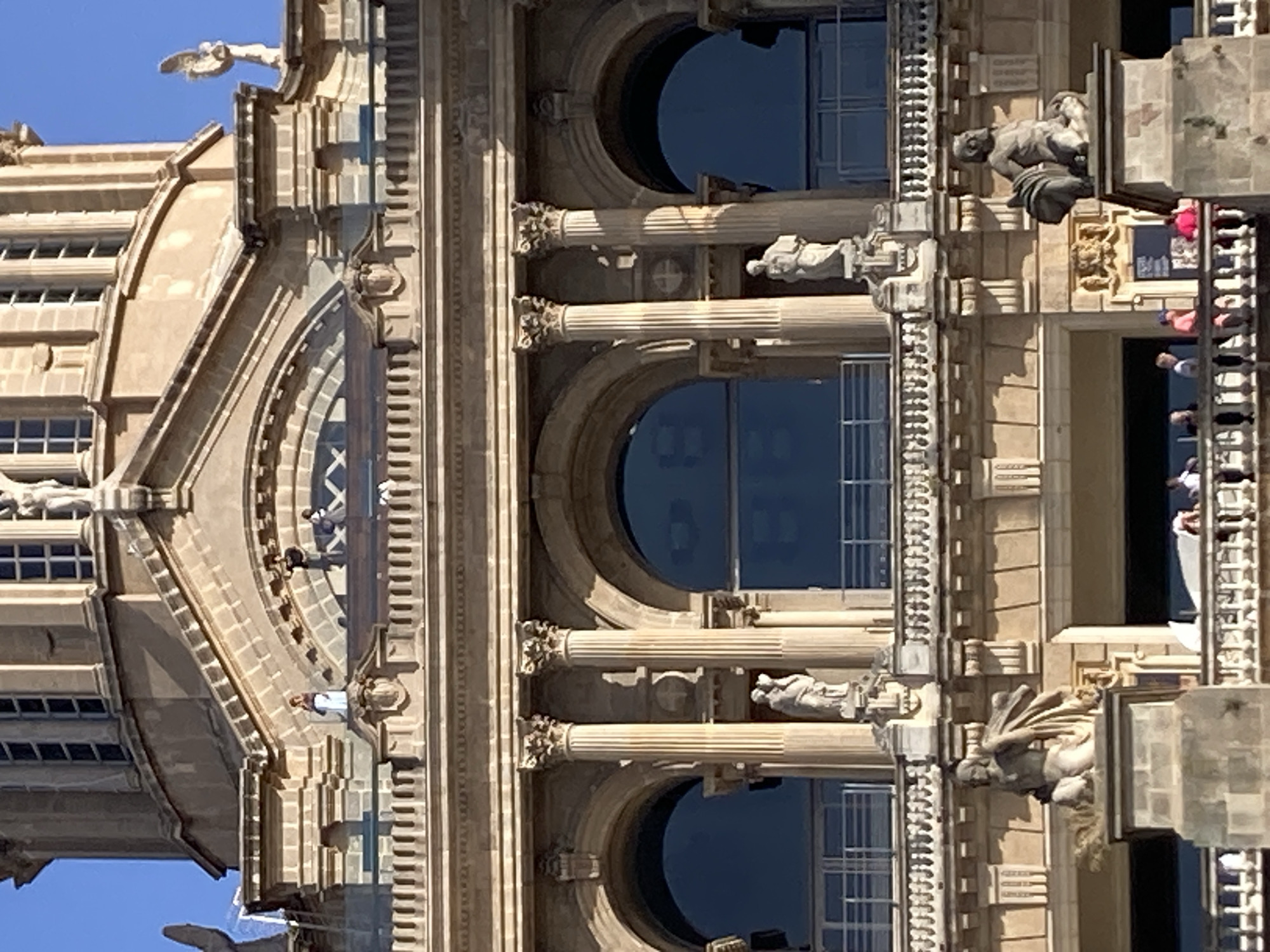 The image shows a close-up, eye-level view of the Palau Nacional, a monumental palace located on Montjuïc hill in Barcelona, Spain. The palace is built in a grand Neoclassical style, characterized by its imposing architecture, arched windows, and ornate balconies. The facade is made of light-colored stone, with decorative elements such as columns, sculptures, and intricate carvings.
In the foreground, a balcony railing is visible with several people standing on it. They appear to be tourists, casually dressed, and observing the surroundings. Further up the facade, on higher balconies and ledges, more people can be seen, some standing and others leaning against railings. These individuals are smaller in the frame, suggesting they are at a greater distance or the photograph is zoomed in on the building's details.
The architectural features are dominant in the image. Two large arched windows with dark blue glass are prominent on the middle level, framed by classical columns. Above these, another level features smaller arched windows and decorative detailing. The top of the image shows a curved dome structure, partially obscured by scaffolding or construction elements, with a bright blue sky in the background.
Several sculptures are integrated into the building's design. Two notable statues stand on plinths on the lower part of the visible facade, one on the left depicting a male figure with draped clothing and another on the right showing a seated male figure with a beard. Smaller statues are also visible on higher levels, integrated into the architectural ornamentation. The overall impression is one of grandeur and historical significance, with the presence of people adding a sense of life and activity to the monumental structure.