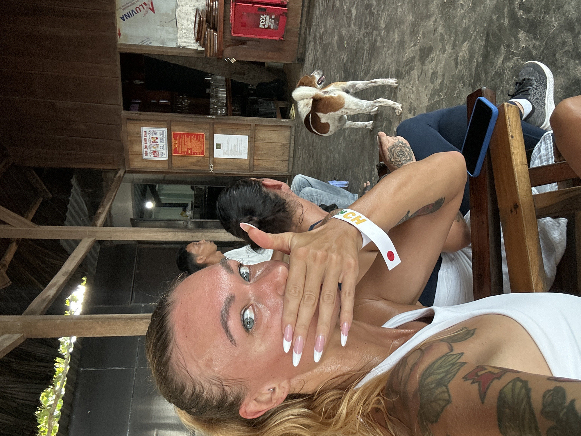 In Mũi Né, Vietnam, a woman with blonde hair in a ponytail, wearing a white tank top and dark leggings, is captured in a close-up selfie. Her right hand covers her mouth, and she wears a white wristband with red and white accents. Her left arm is visible, displaying prominent tattoos. A blue smartphone rests on the wooden chair beside her. In the background, a brown and white dog stands on a concrete floor. Two other individuals are partially visible, one in a white shirt and another with dark hair. The environment appears to be an open-air structure with a thatched roof and wooden walls. A sign with Vietnamese text is mounted on a wooden door.