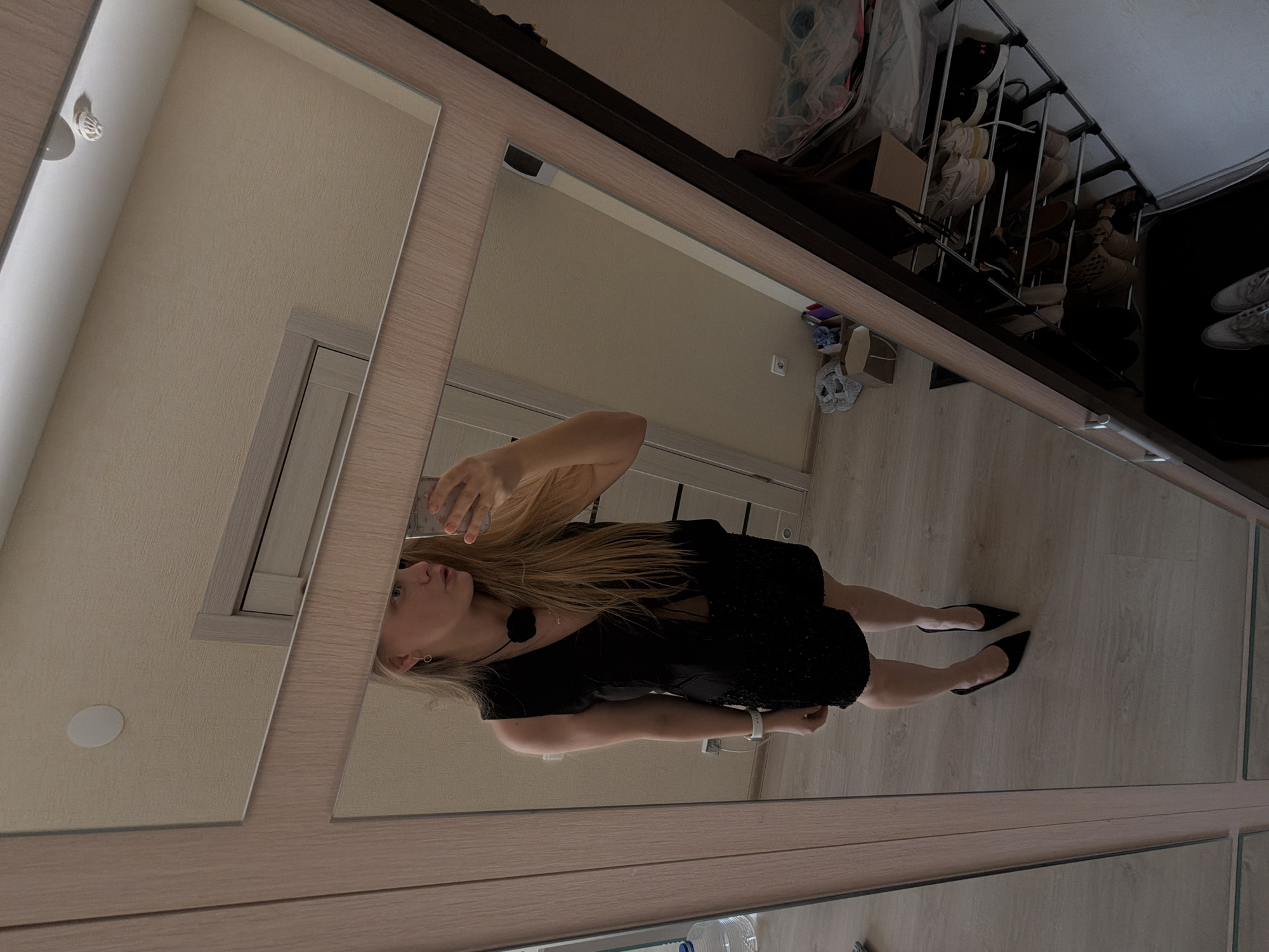 A woman is seen in the reflection of a large mirrored wardrobe. She has long blonde hair and is wearing a short black dress and black high heels. She is holding a phone in her left hand, presumably taking the photo. The wardrobe has a light-wood frame with multiple rectangular mirrors. In the background, partially visible above the wardrobe, is a shoe rack holding various pairs of shoes. The surrounding environment seems to be an indoor space, most likely a bedroom or dressing room. There is no information that would suggest that the image was taken in Izhevsk, Russia.