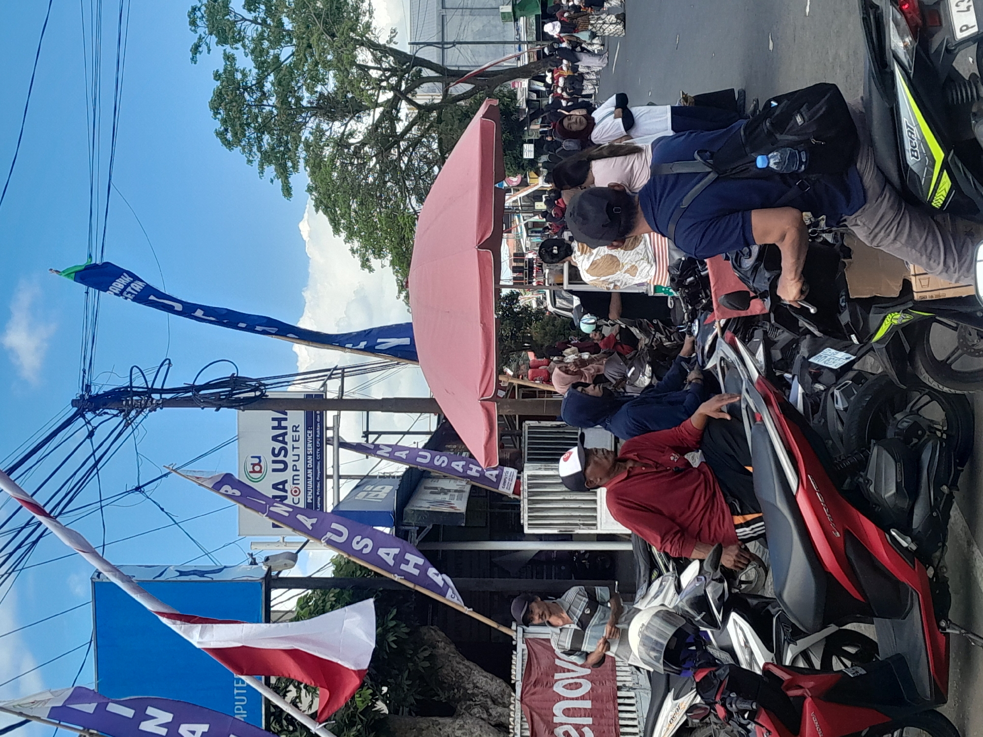 In Genteng Kulon, Indonesia, an outdoor scene depicts a street lined with shops and filled with people and motorcycles. Flags, including Indonesian national flags and purple banners with text like "NA USAHA COMPUTER," are visible, suggesting a local event or business promotion. Several individuals are seen on and around parked motorcycles. One man in a red shirt and black cap is seated on a red motorcycle. Another man in a blue shirt and a black backpack is seated on a black motorcycle, with a water bottle attached to his bag. A group of people, some in traditional attire, are gathered further down the street. A large pink umbrella provides shade over a stall. The sky is partly cloudy, and trees are present in the background. The overall impression is a bustling street scene with commercial activity and community gathering.