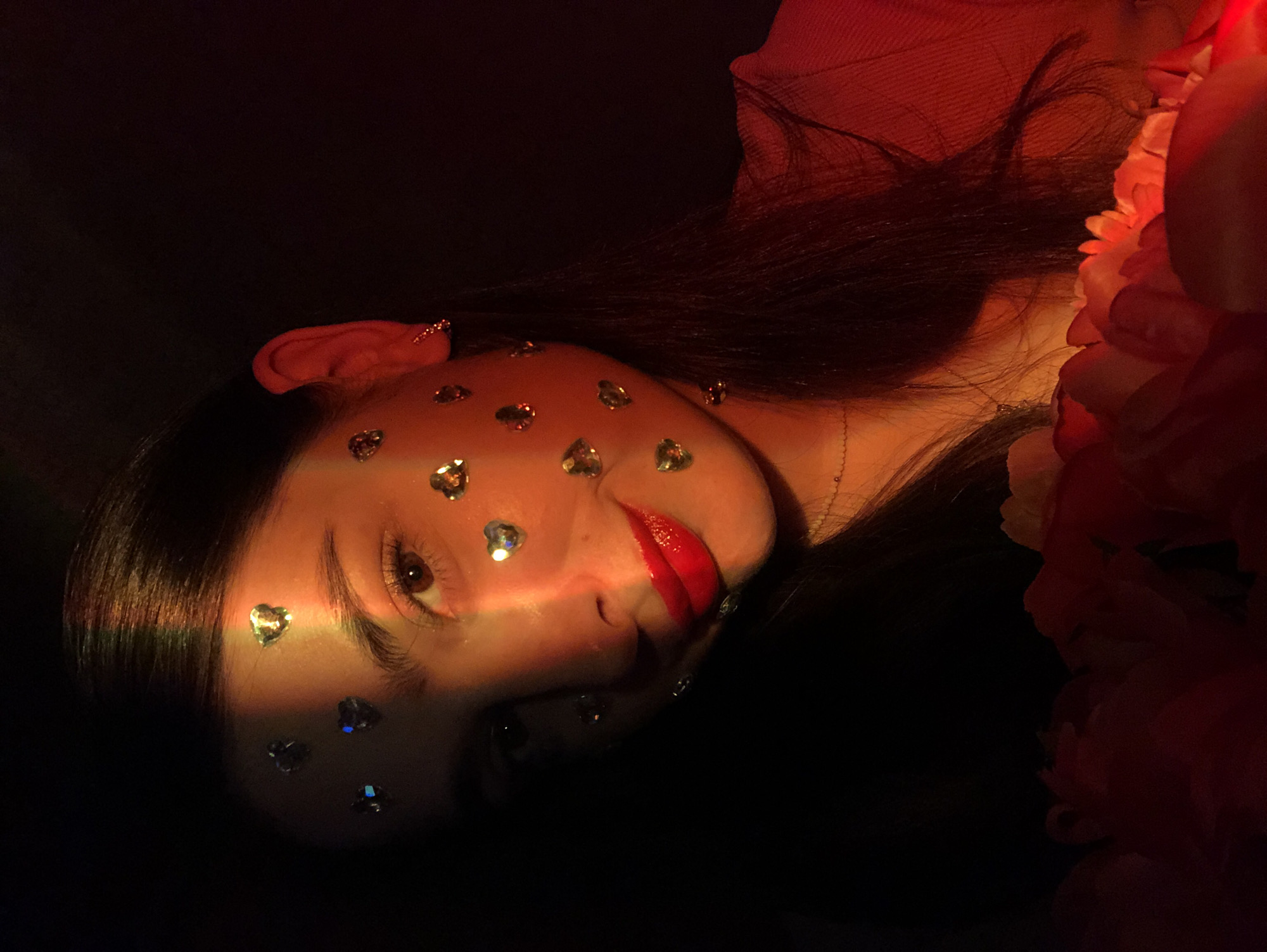 The image shows a close-up of a young woman's face, adorned with heart-shaped sequins. She has dark hair, brown eyes, and is wearing bright red lipstick. A colorful light spectrum, possibly from a prism or colored lighting, casts a shadow across her forehead and cheek. The background is dark, with a hint of red lighting on the right side. In the foreground, there are red flowers. The scene appears to be indoors, possibly a dimly lit room, with no discernible elements placing it specifically in Noviy, Russia.