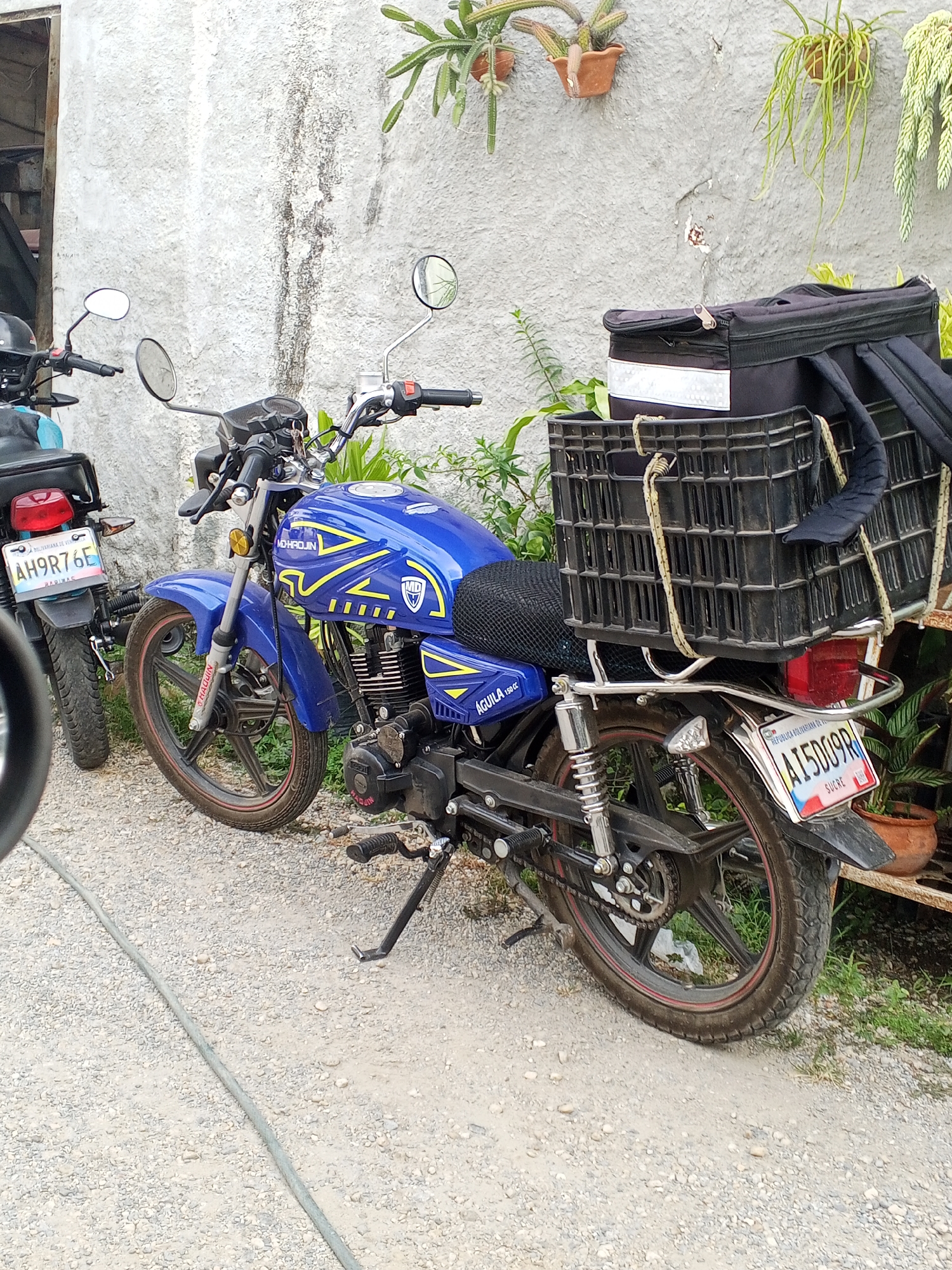 The image depicts two motorcycles parked on a gravel surface. The one in the foreground is blue and has a black storage box attached to the back. A black crate is also attached to the back of the motorcycle with what appears to be rope. The license plate is visible and reads "A15D09R" with the word "SUCRE" underneath it, suggesting it's from the Sucre state in Venezuela. The other motorcycle is dark green and only partially visible in the background; its license plate reads "AH9R76E". The motorcycles are parked next to a weathered white wall with some plants hanging from it. Based on the license plate, the location is likely in Venezuela. The city is not directly evident from the image.