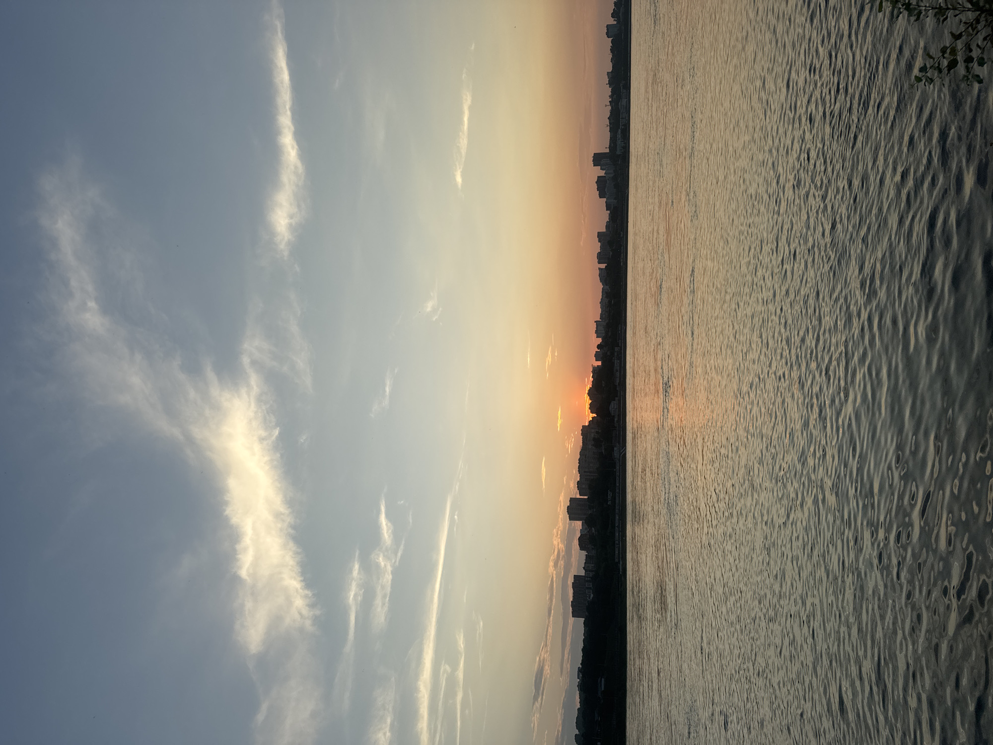 The image captures a sunset over the Angara River in Irkutsk, Russia. The sky is a gradient of blue and orange, with wispy white clouds. In the distance, a city skyline is visible, with buildings of various heights silhouetted against the colorful sky. The river in the foreground is calm, reflecting the colors of the sunset. The overall impression is one of a peaceful and serene evening in the city.