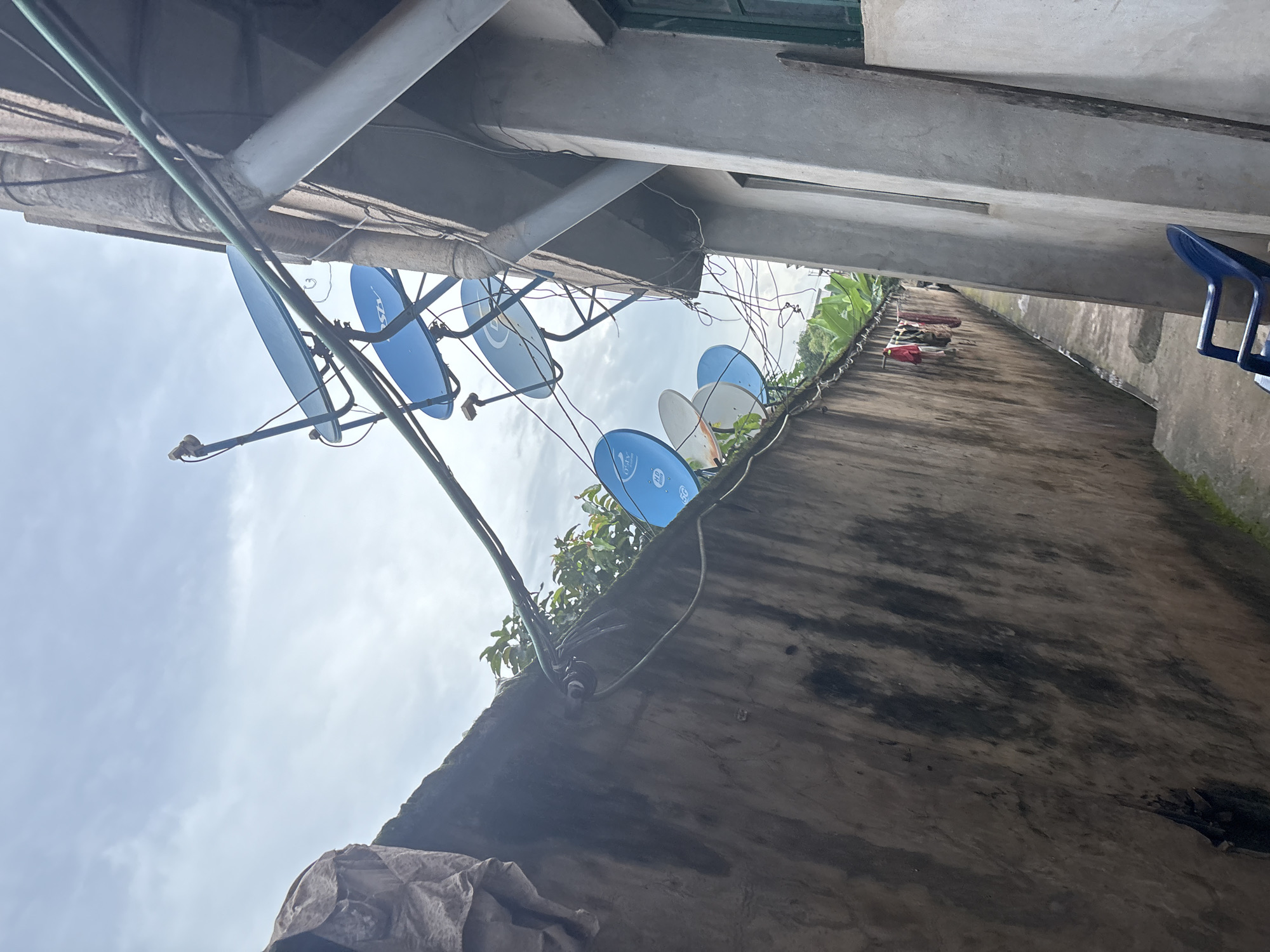 The image depicts an outdoor scene in Onitsha, Nigeria, featuring a narrow alleyway between two buildings. On the left, a weathered, concrete wall runs horizontally across the frame, covered in grime and dark stains, with a piece of brown tarp draped over its top left corner. Several satellite dishes, primarily blue and white, are mounted on poles and wires that extend from the wall and a nearby building on the right. The building on the right is a rough, concrete structure with a green window visible and a large, smooth concrete pipe running vertically. Wires are strung haphazardly around the satellite dishes and between the buildings. In the alley, there is a small patch of green vegetation, and clothes are hanging on a line attached to the left wall. A blue plastic chair is situated in the foreground on the right side of the alley, near the concrete building. The sky above is overcast.