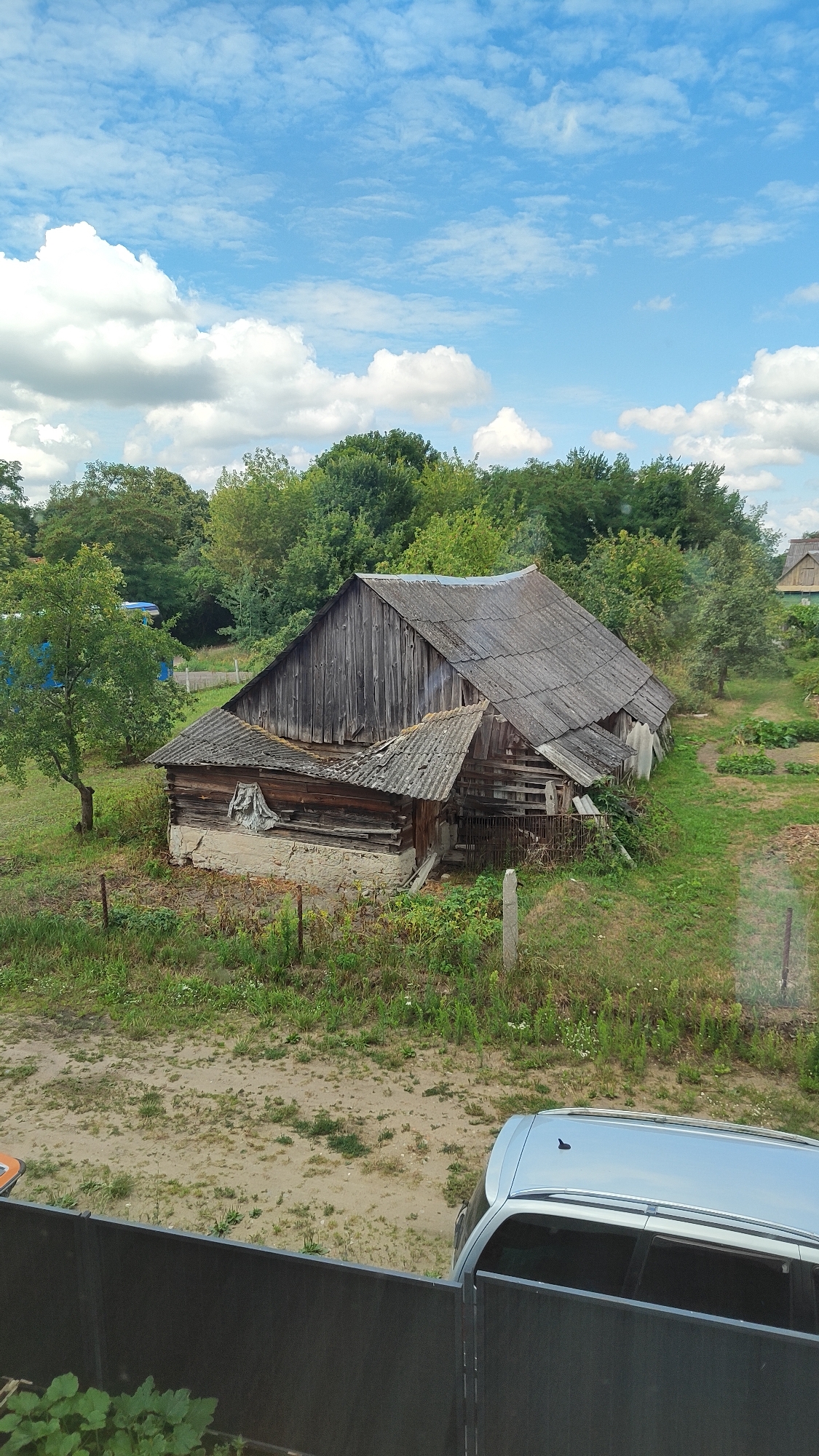 An exterior shot shows a dilapidated wooden barn with a tin roof, surrounded by lush green trees and overgrown grass under a bright blue sky with scattered white clouds. In the foreground, a silver car is parked behind a dark fence. The scene suggests a rural or semi-rural setting, possibly in the Тэльмінскі сельскі Савет region of Belarus, given the architectural style and vegetation. The barn appears to be old and in disrepair, with weathered wooden planks and a partially collapsed roof. The surrounding area is dotted with more trees and some evidence of cultivated land, further reinforcing the impression of a rural environment. The overall mood is one of quiet abandonment and the passage of time.