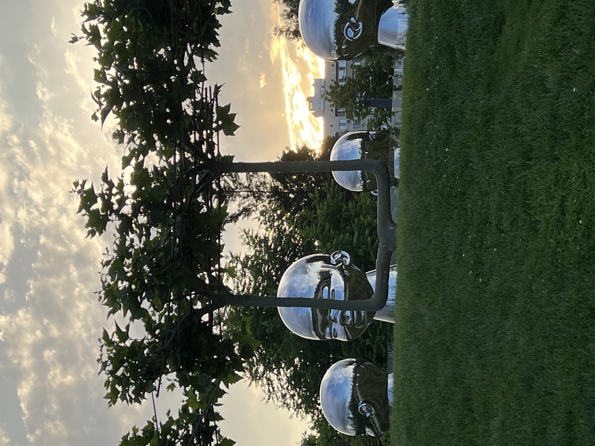 In Krasnodar, Russia, at the Park of the First World War, a group of mirrored sculptures of giant baby heads are displayed on a grassy knoll. The sculptures are part of a public art installation. In the background, trees and a building can be seen. The sky is overcast with the sun peeking through the clouds. The sculptures are reflective, mirroring the sky and surrounding environment. The overall scene is serene and somewhat surreal.