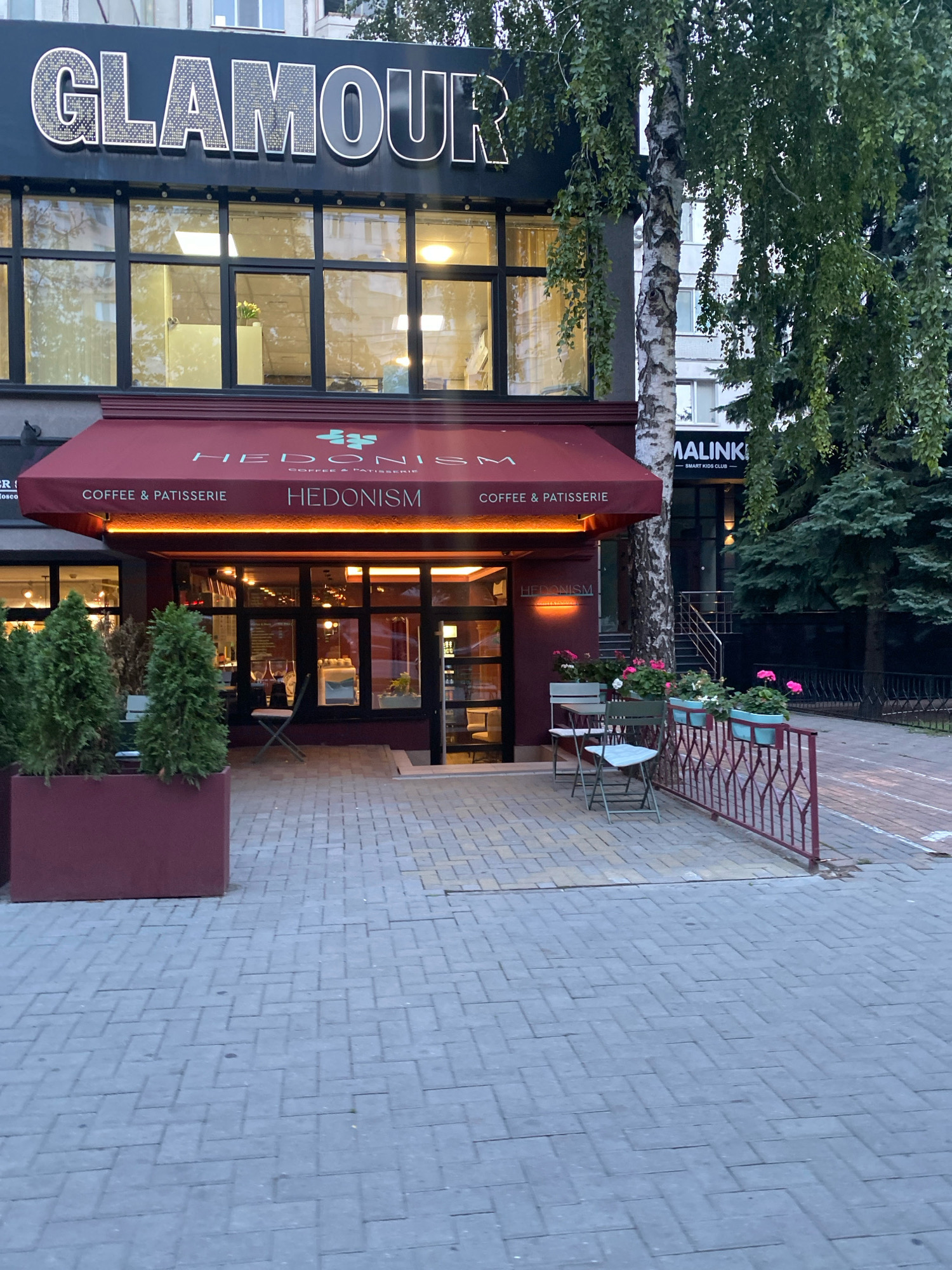 The image shows a street-level view of the exterior of a "GLAMOUR" building and a "HEDONISM COFFEE & PATISSERIE" establishment in Chișinău, Moldova. The "GLAMOUR" signage is prominent at the top of the building. Below it, large windows reflect the interior lights. A dark red awning with "HEDONISM" and "COFFEE & PATISSERIE" written on it covers the entrance to the cafe. The cafe has a glass storefront, allowing a view into the interior. Outside the cafe, there are two small tables with chairs, a red metal railing, and planters with flowers and small evergreen trees. In the background, a birch tree stands next to another building with signage that includes "MALINKI - SMART KIDS CLUB". The ground in front of the establishments is paved with grey rectangular bricks.