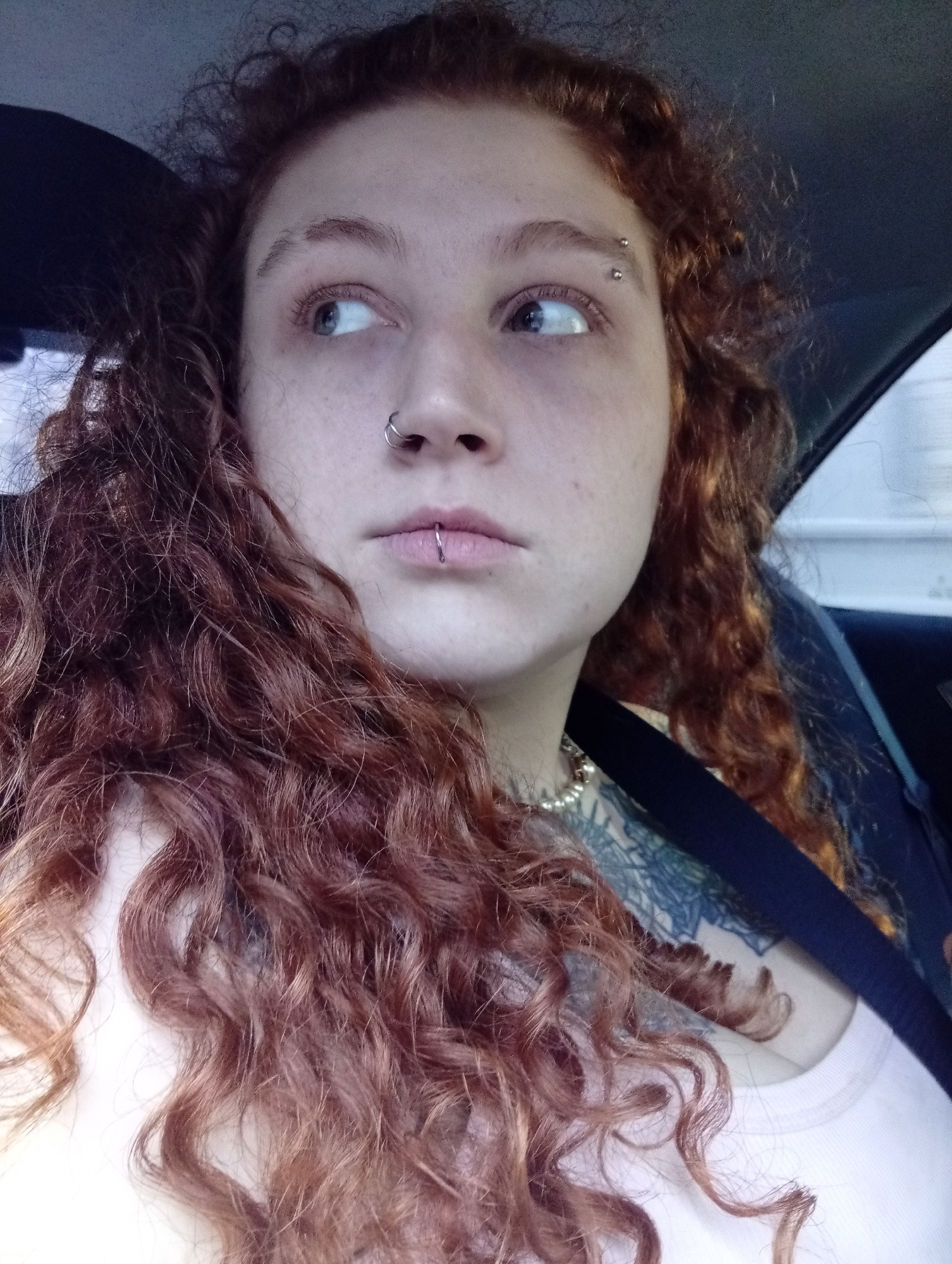 A young person with red curly hair, an eyebrow piercing, a nose ring, and a lip ring is seated in what appears to be a car. The person is wearing a seatbelt and a light-colored top. Part of a tattoo is visible on the person's upper chest. The interior of the car is visible, along with a hint of a building outside the window. No information suggests the location is Buenos Aires, Argentina.