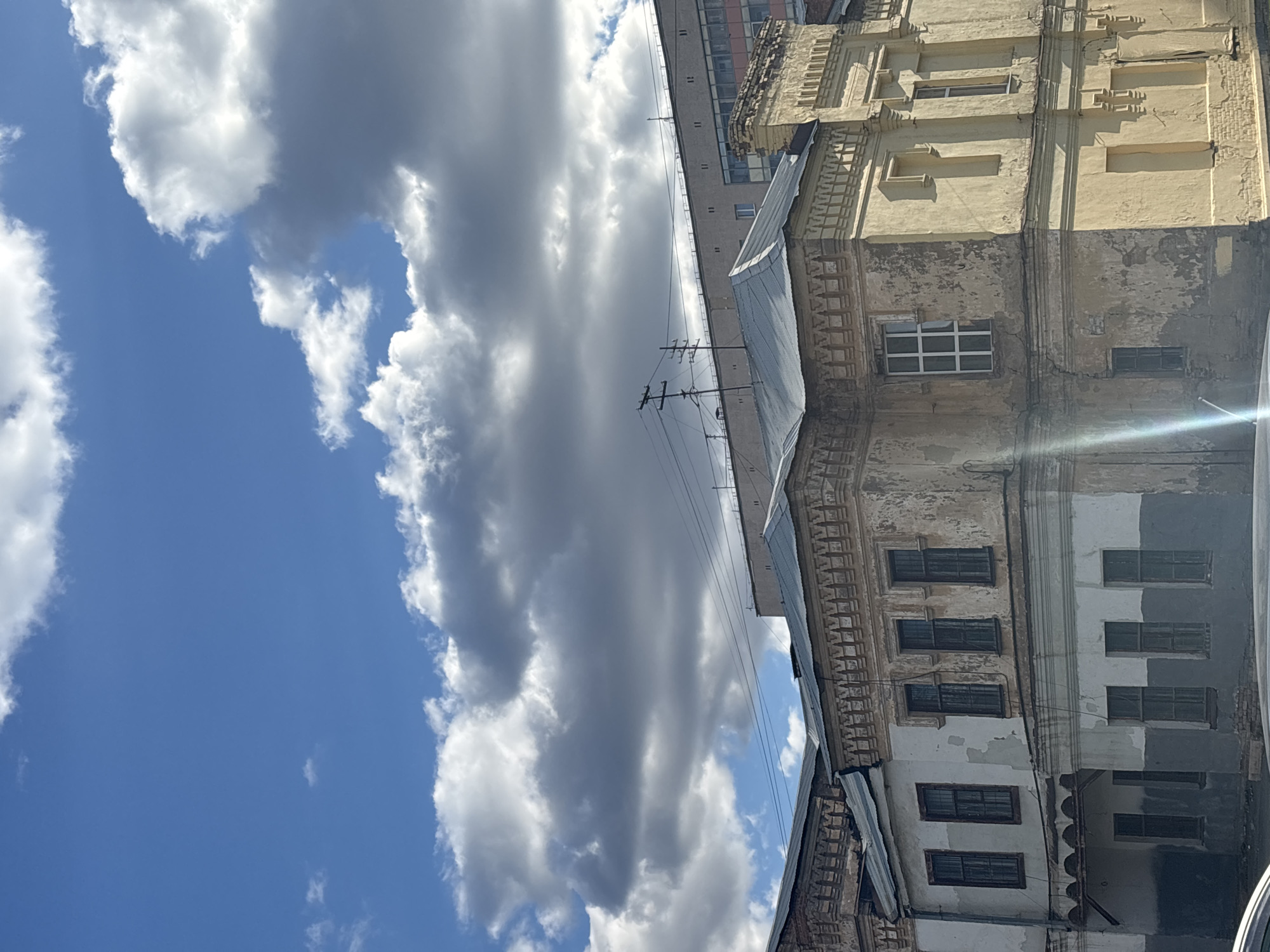 The image shows a view of Orenburg, Russia. A building with a yellow facade and visible signs of age stands in the right portion of the frame. The building has multiple windows and architectural details. Overhead electric wires are visible. A sky filled with white and gray clouds dominates the left side of the image. The sky transitions from blue to white as the clouds take over.