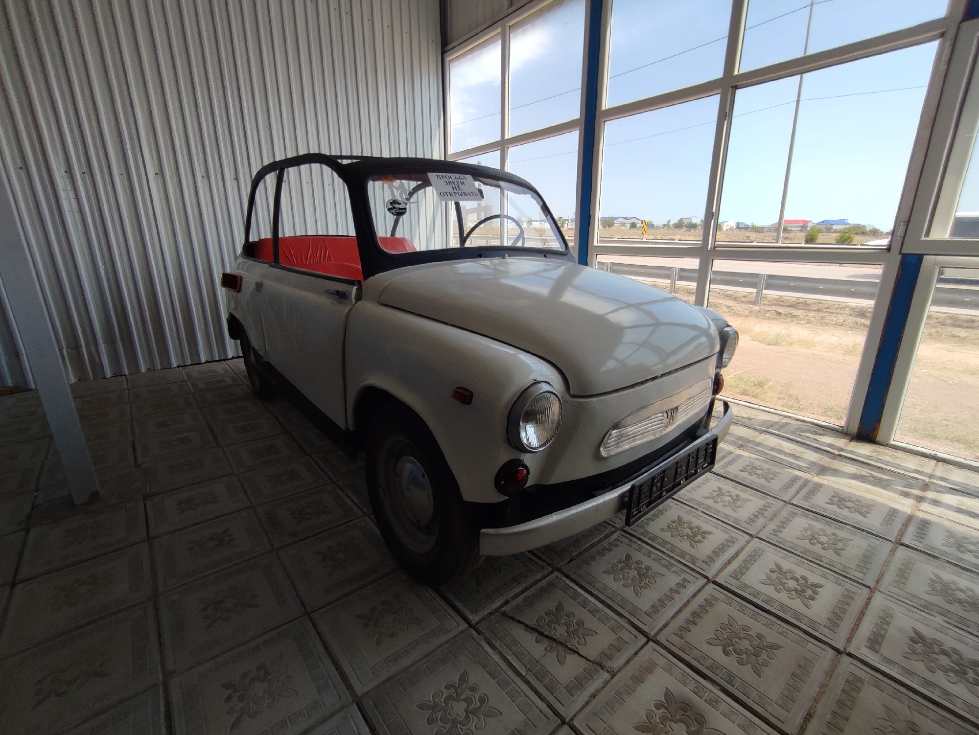 An unidentifiable white, convertible microcar is parked indoors on a tiled floor. The car has a black soft-top frame and red interior seats. A sign on the windshield reads "ПРОСЬБА ДВЕРИ НЕ ОТКРЫВАТЬ" which translates to "PLEASE DO NOT OPEN THE DOORS." The car is positioned in front of a large window that looks out onto a road with houses and buildings in the distance. The setting appears to be a museum or exhibition space located in the city of Qonaev, Kazakhstan, a location known for its unique architecture and proximity to the Kapchagay Reservoir. The overall scene is well-lit, with natural light streaming in from the windows, casting shadows on the floor and the car.