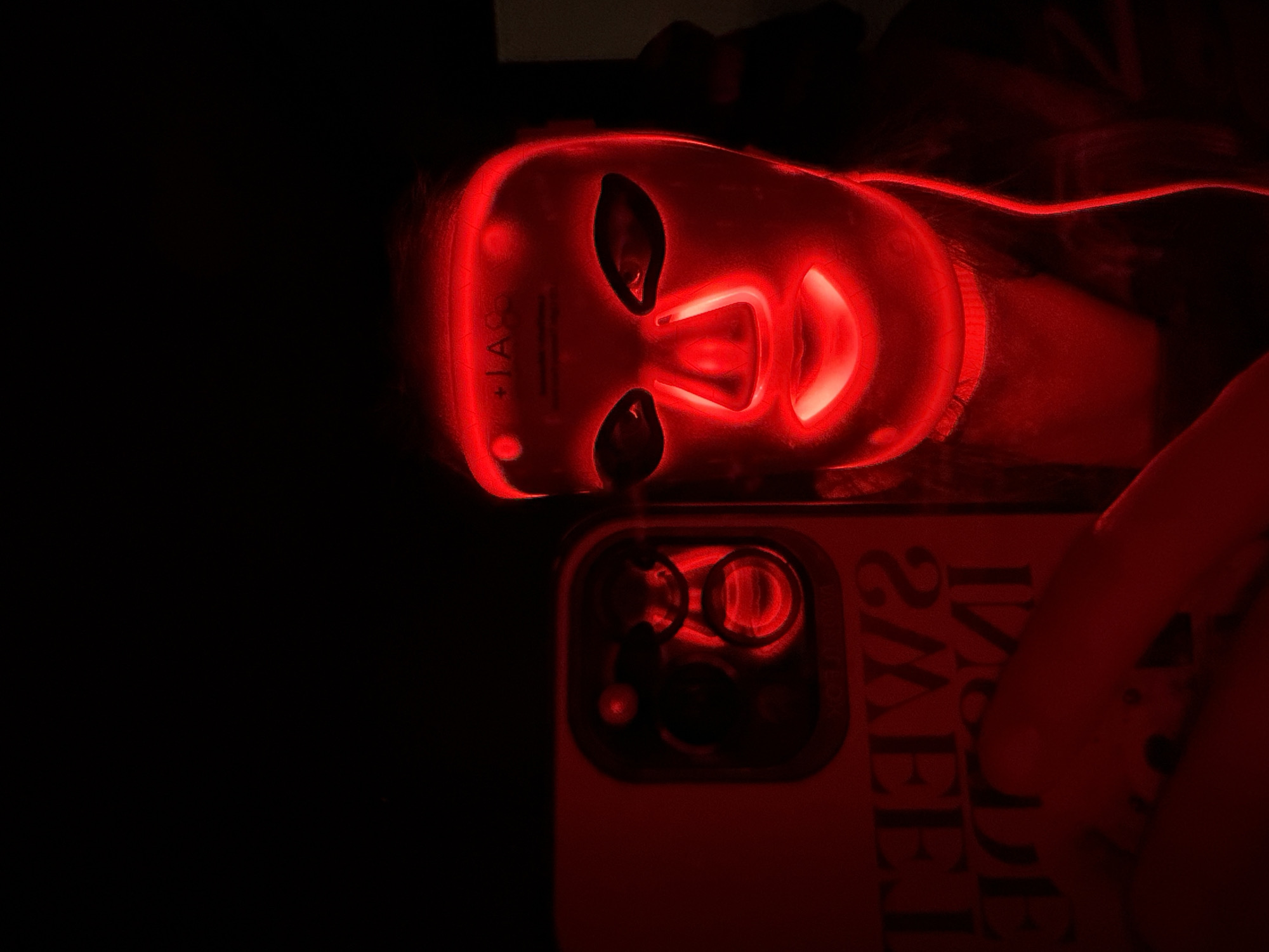 A person is seen in a dark setting, lying down, with their head illuminated by a bright red light emanating from a facial mask. The mask, a red light therapy device, covers the entire face with cutouts for the eyes, nose, and mouth, and glows intensely. Text resembling "IAC" or "IAG" is visible on the left side of the mask. The person's hair is dark, and their face is obscured by the glowing mask.
To the right and below the person's head, a smartphone is visible, resting on a surface. The back of the phone is encased in a reddish cover, and its multi-lens camera module reflects the red light from the mask. Text is partially visible on the phone case, appearing upside down and mirrored due to the angle, suggesting phrases like "SWEET" or "2WEE7". The overall scene is dark, suggesting an indoor setting, likely in the evening or at night. The activity taking place is red light therapy, a skincare treatment. The mood is somewhat futuristic or focused on personal care.