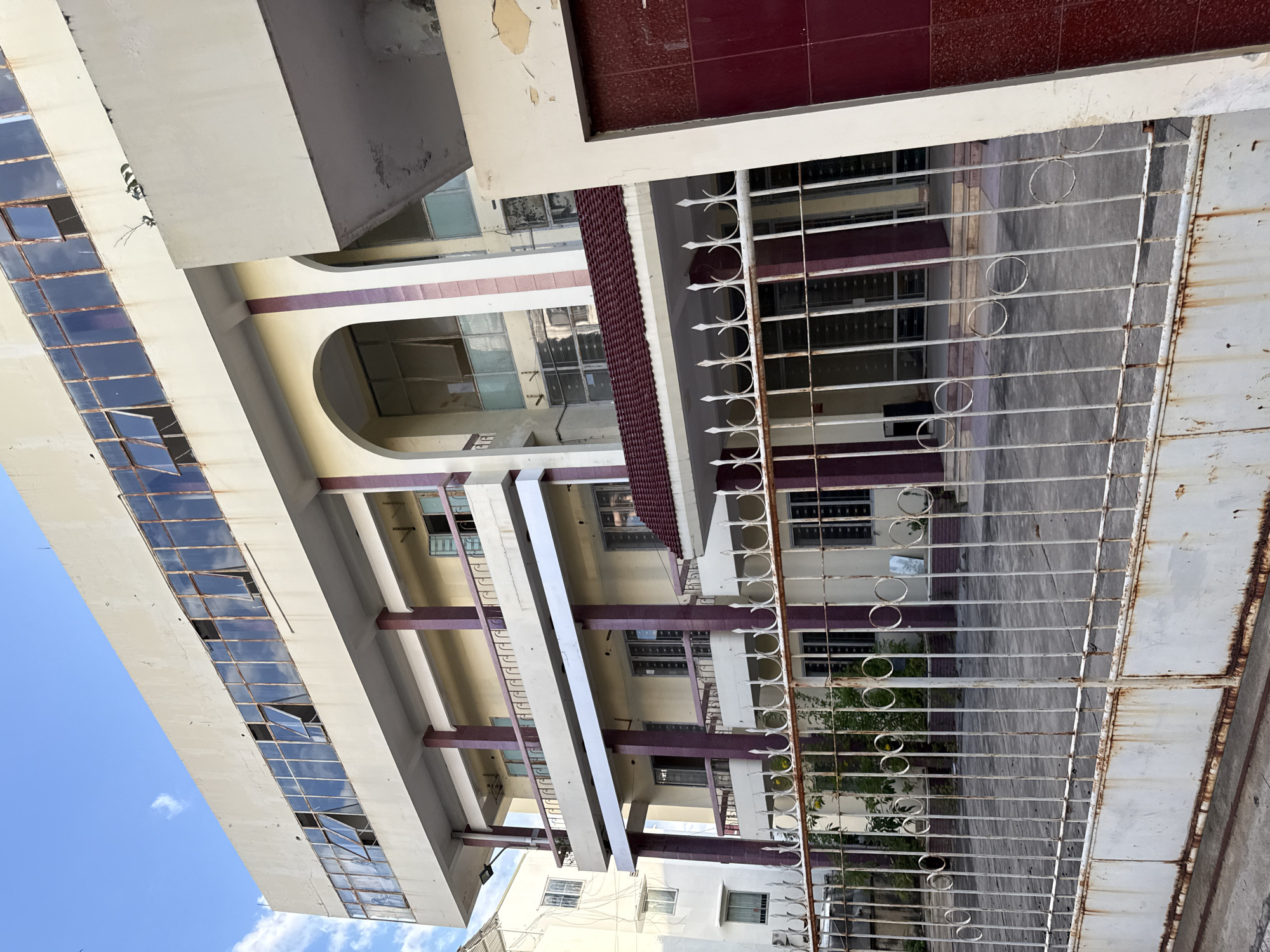 The image depicts the exterior of a multi-story building in Nha Trang, Vietnam, with a focus on its facade and a foreground gate. The building, likely an office or institutional structure, exhibits a weathered appearance. Its upper floors feature a long row of windows with metal frames, some of which are open, revealing reflections of the sky. The walls are painted a light cream color, with decorative purple columns supporting the upper levels and balconies. The building has arched entrances on the lower floors and a red tiled roof section over one entrance. A white metal gate, showing signs of rust, stands in front of the building, with vertical bars and circular decorative elements. The ground in front of the gate appears to be paved. Green foliage is visible behind the gate, suggesting some landscaping. The sky is blue with scattered white clouds. The overall impression is one of a slightly neglected but functional urban building.
