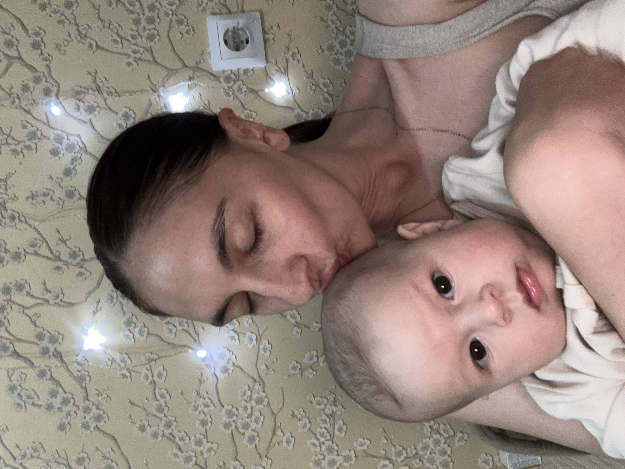 A woman is lying on a surface with a floral pattern, illuminated by small, white lights. She is kissing a baby who is lying on top of her. A white electrical outlet is visible near the woman's head. The background is blurred. The scene does not show anything to suggest that it takes place in Sochi, Russia.